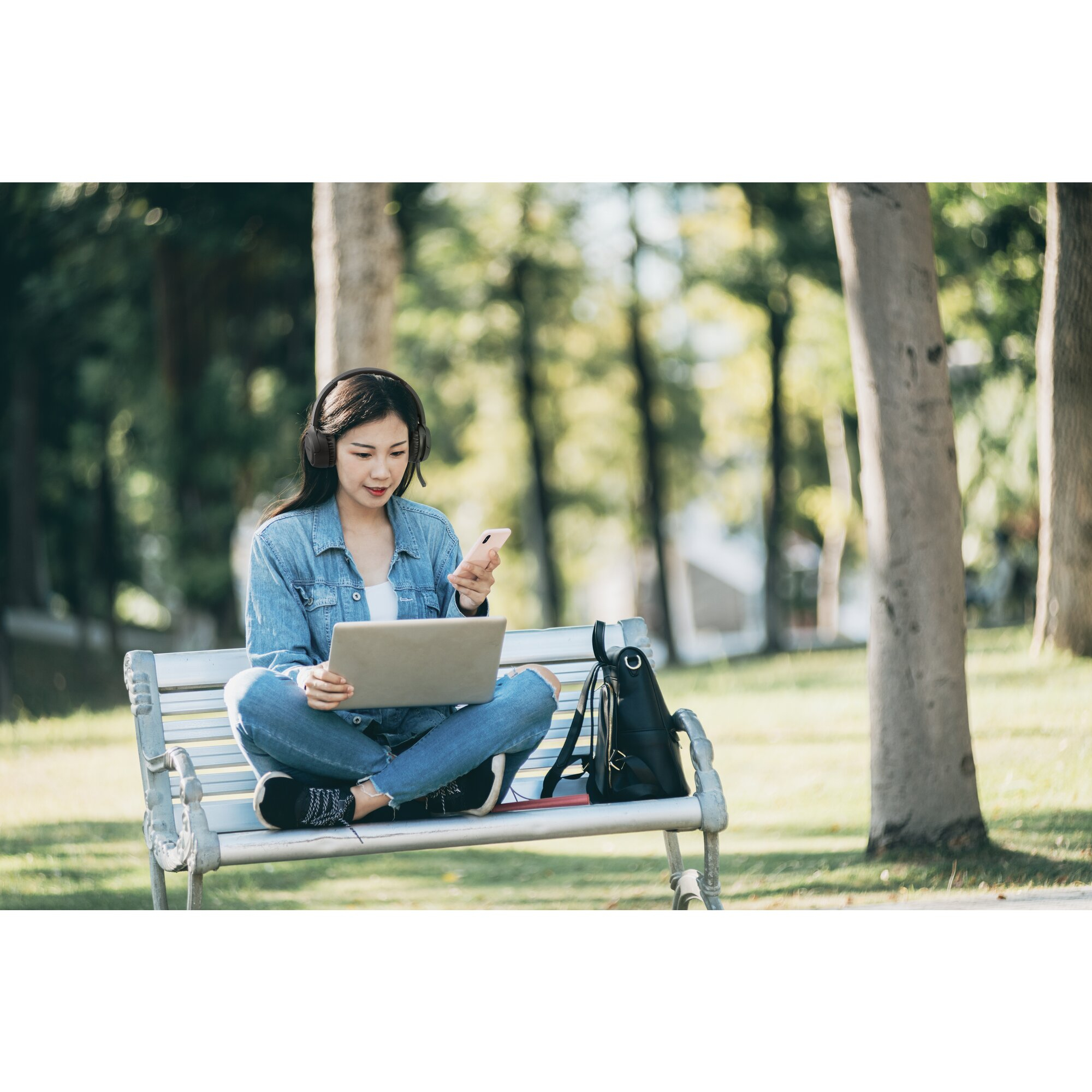 Kobieta siedzi na ławce w parku, używając laptopa i telefonu, ma słuchawki, kurtkę dżinsową i plecak.