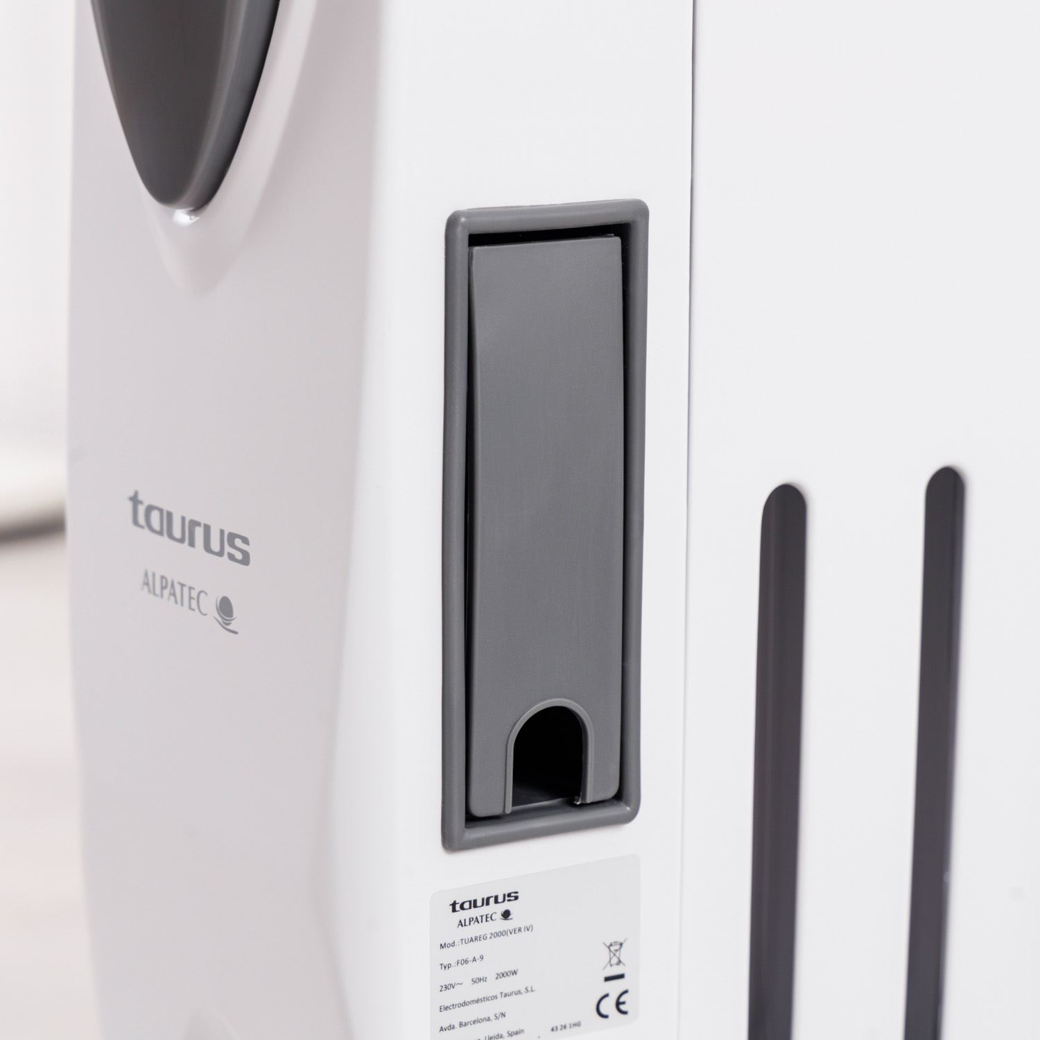 Primer plano de un electrodoméstico blanco con detalles grises. Se ve la marca 'Taurus' y el modelo 'ALPATEC'. También un panel y rejillas grises.