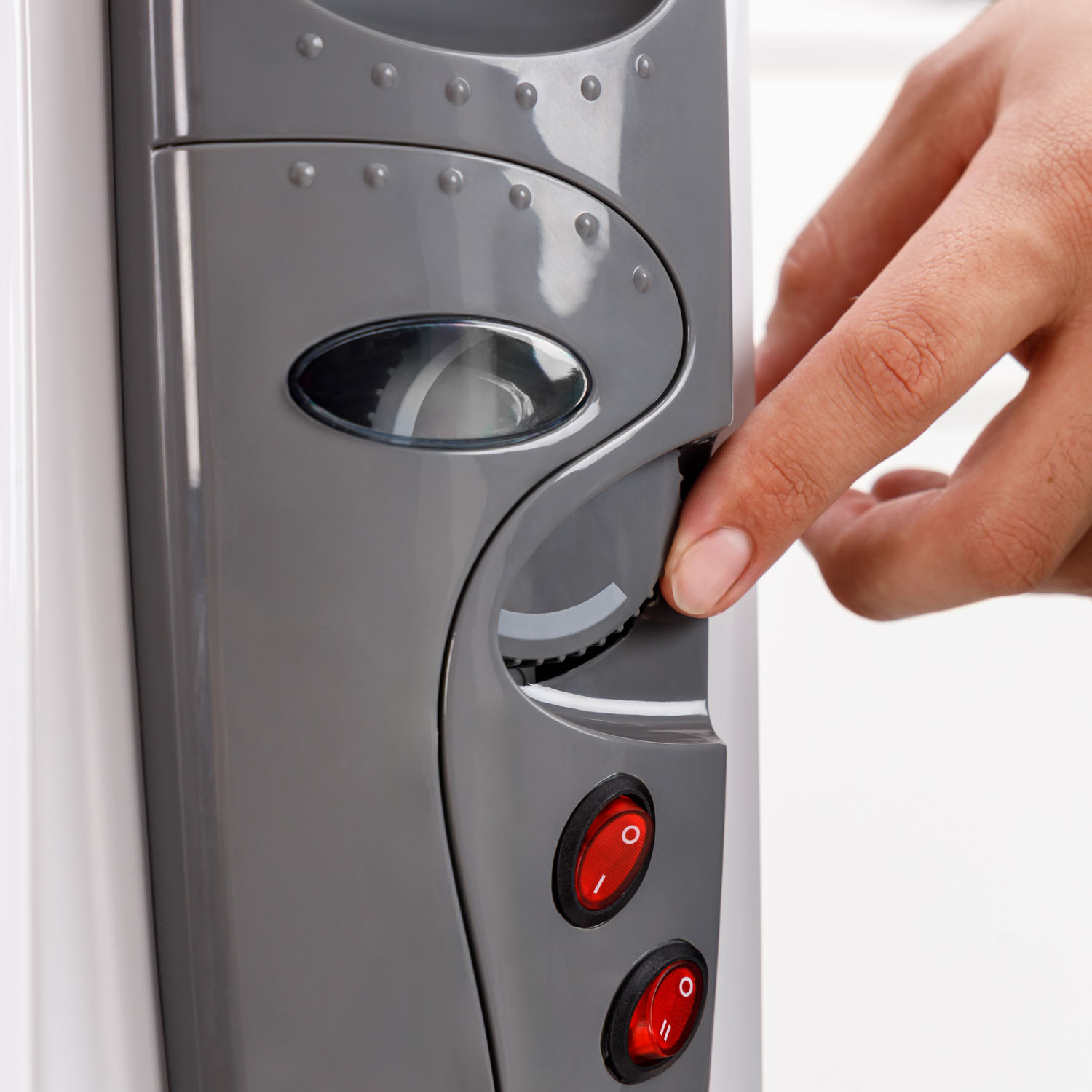 Una mano ajusta el dial en un calefactor eléctrico gris y blanco. Se ven botones rojos de encendido. El fondo es blanco.