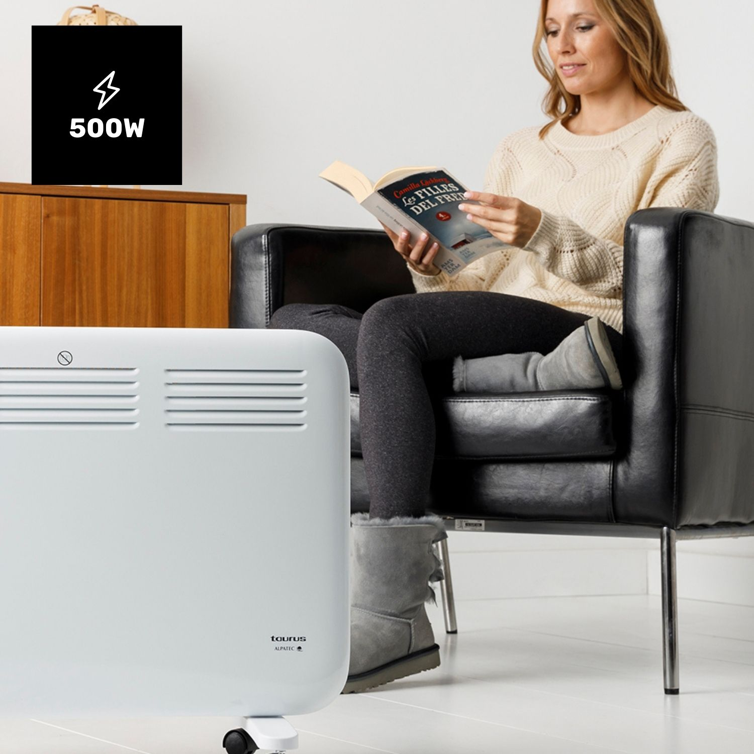 Mujer leyendo un libro, sentada en un sillón negro. Un calefactor blanco está frente a ella. Un letrero de 500W está a la izquierda.