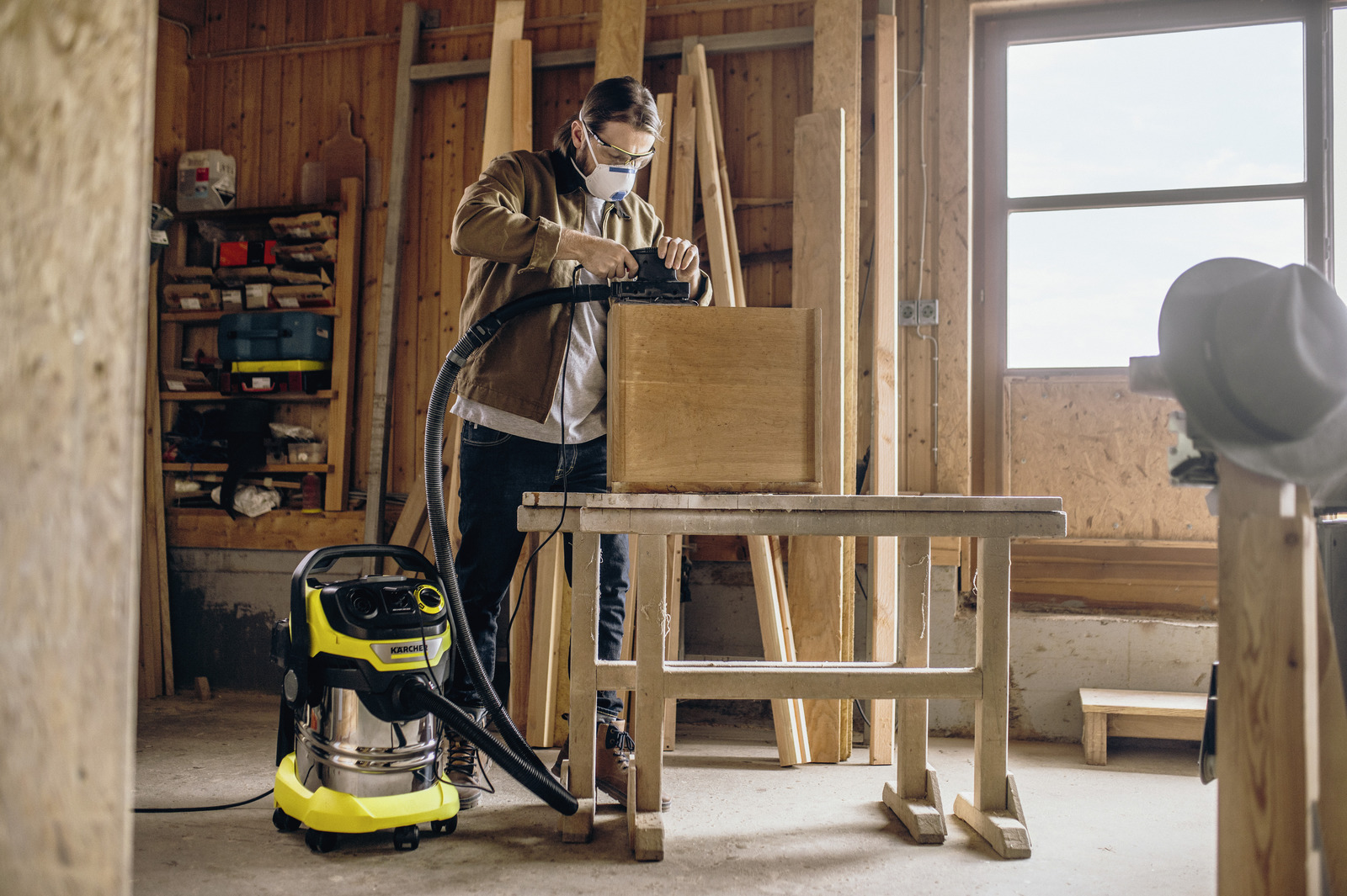 Ein Mann schleift in einer Werkstatt eine Holzkiste. Ein Staubsauger steht daneben. Der Raum hat Holzwände und ein Fenster.