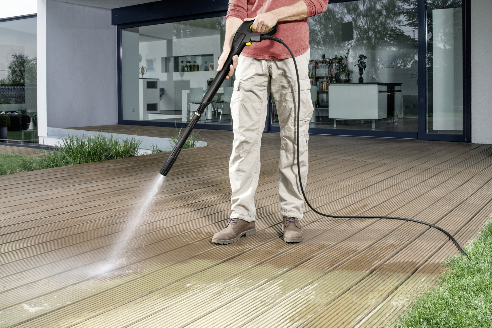 Una persona utiliza una hidrolavadora para limpiar una terraza de madera. El agua sale a chorros de la boquilla, limpiando la madera. El fondo muestra una casa.