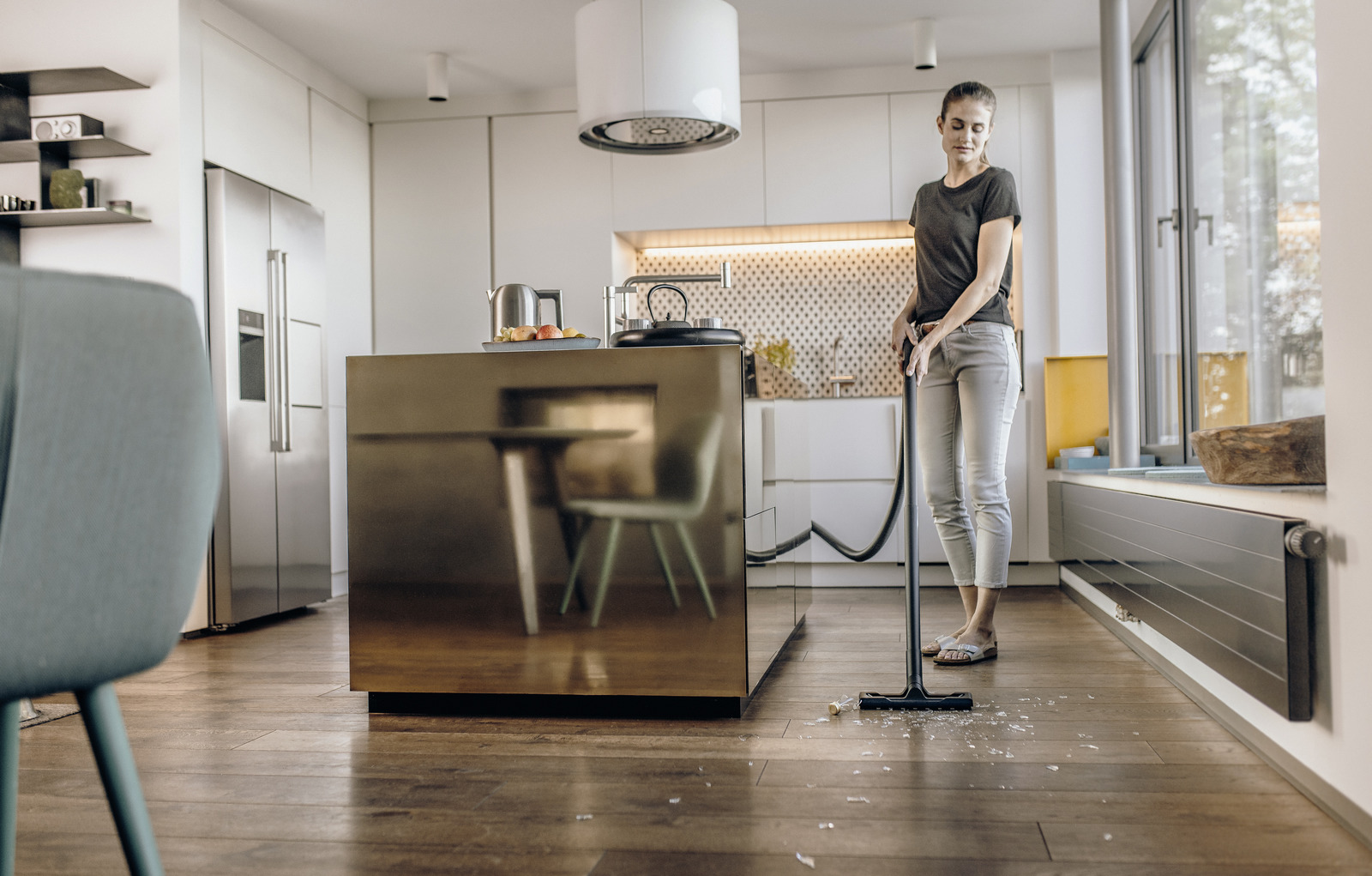 Kobieta odkurza podłogę w kuchni. Kuchnia ma dużą wyspę. Na podłodze są śmieci. Widoczna jest lodówka.