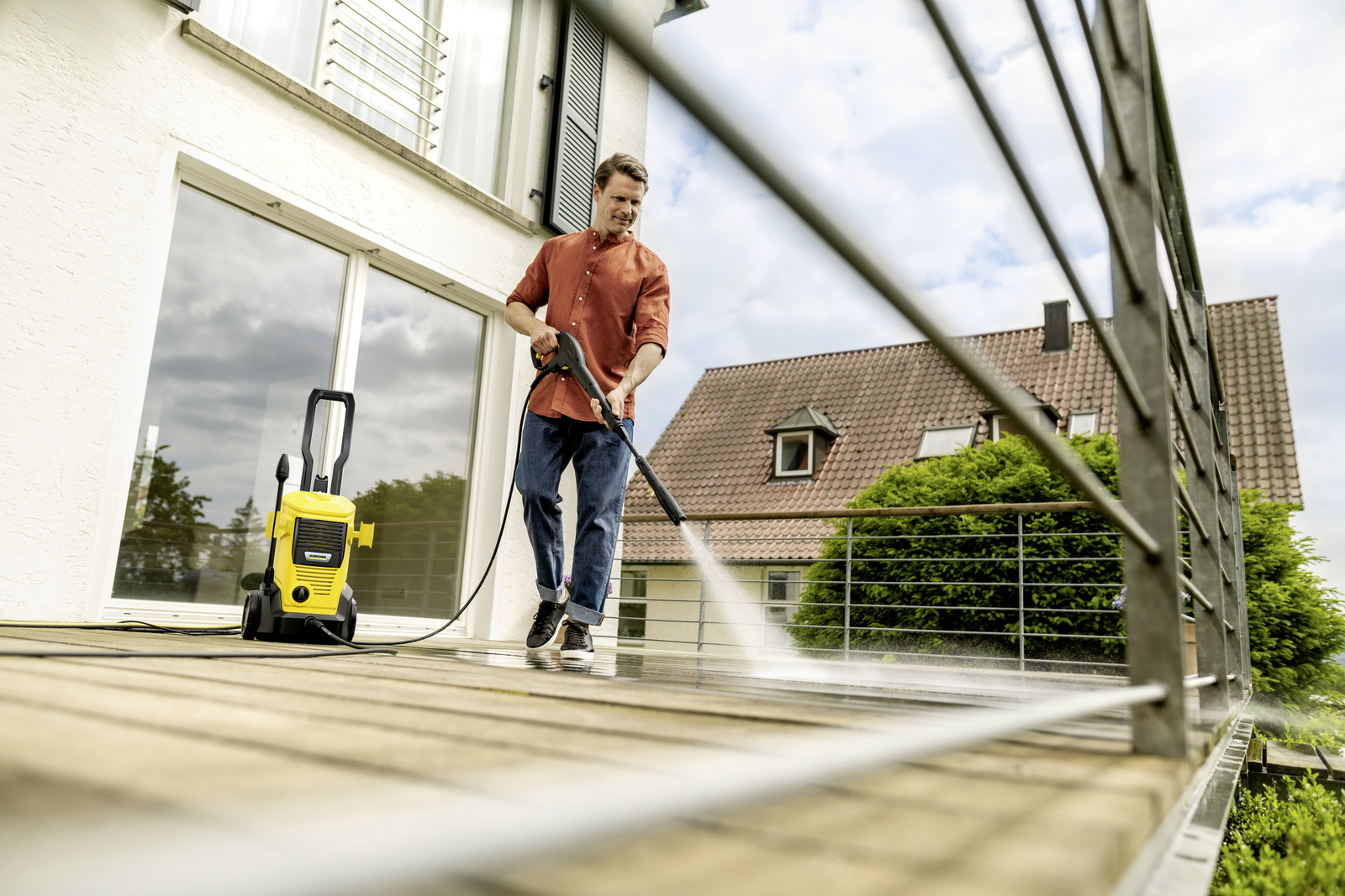 Ein Mann reinigt eine Holzterrasse mit einem gelben Hochdruckreiniger. Ein Haus und Bäume sind im Hintergrund. Der Himmel ist sichtbar.