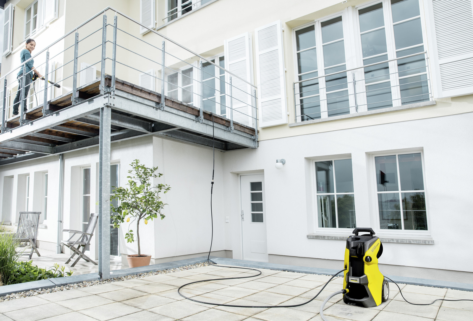 Eine Frau benutzt einen Hochdruckreiniger auf einem Balkon. Ein gelb-schwarzer Hochdruckreiniger steht auf der Terrasse. Weißes Gebäude mit Fenstern.