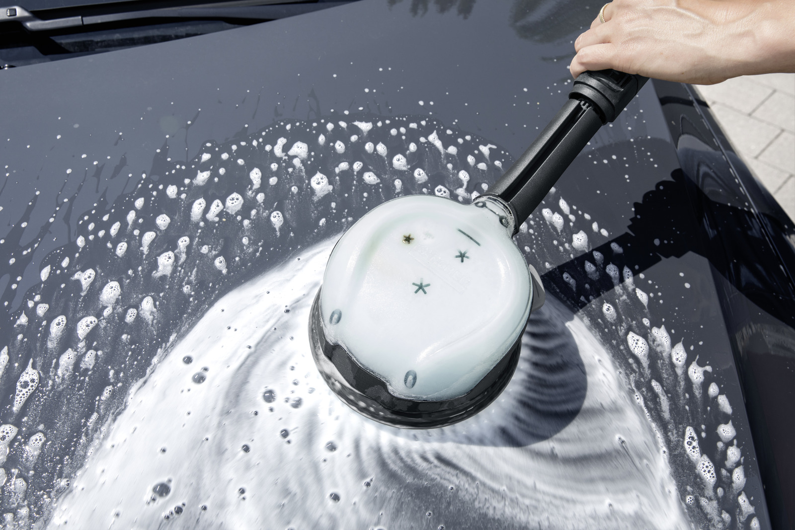 Eine Hand hält eine Autowaschbürste mit Schaum auf einer dunklen Motorhaube. Die Bürste wird zum Reinigen verwendet.