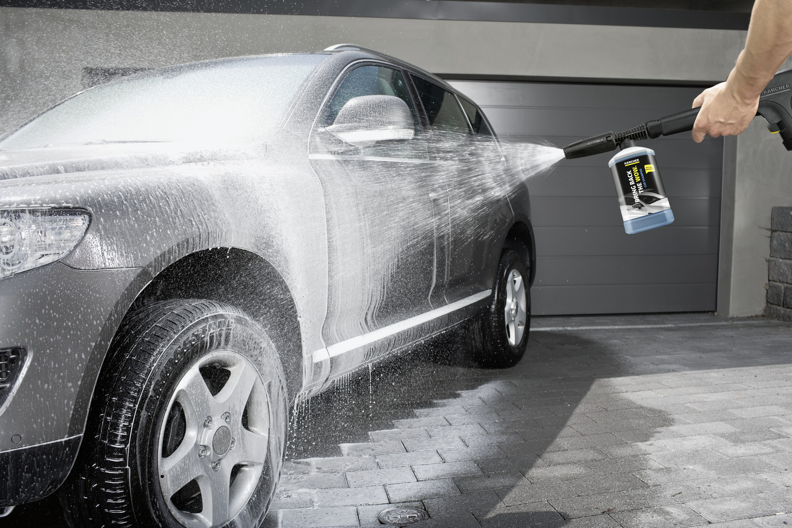 Ein graues Auto wird mit einem Hochdruckreiniger gewaschen. Wasser und Seife besprühen das Auto. Eine Flasche Autowaschseife ist zu sehen.