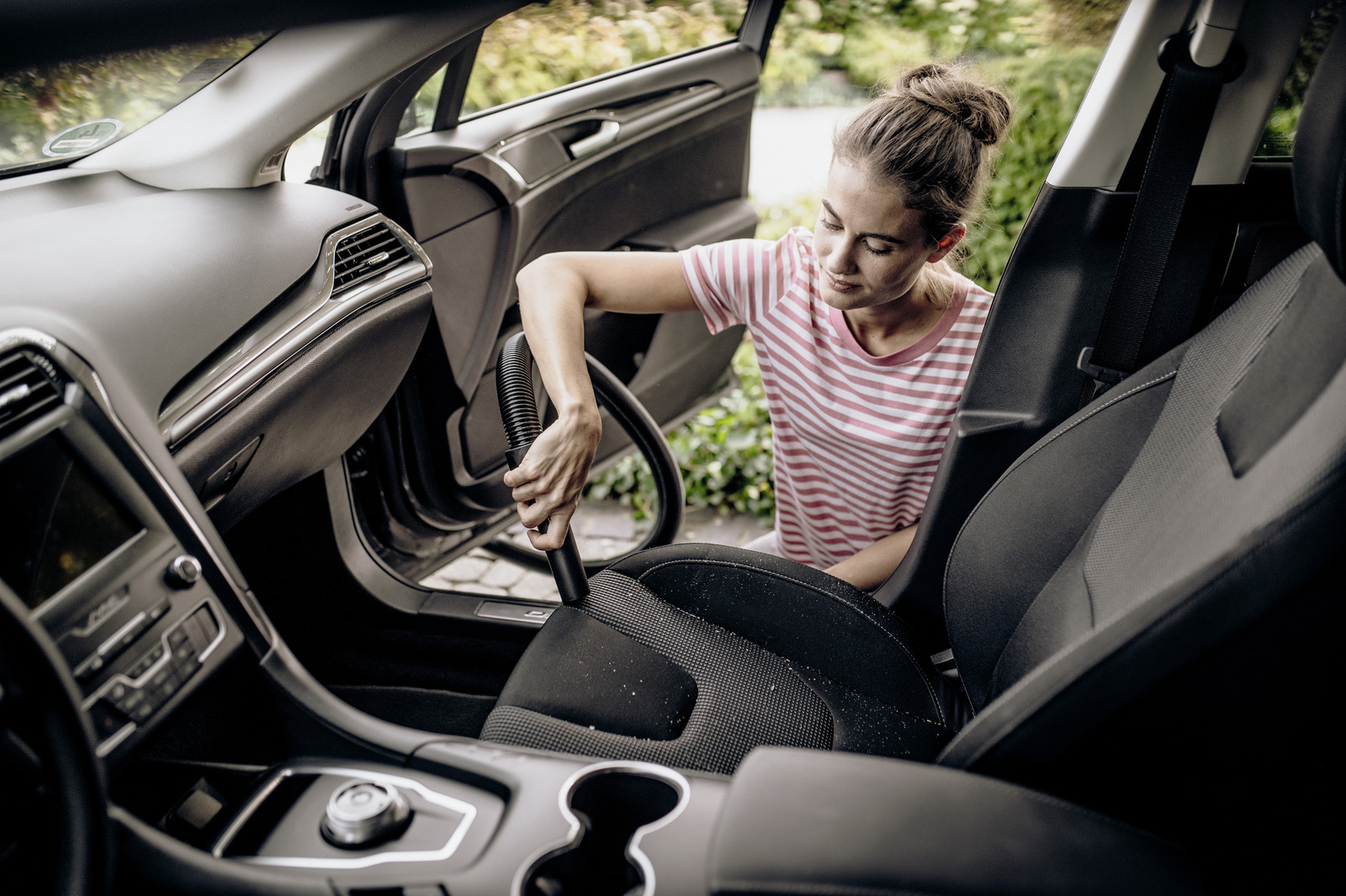 Una donna aspira l'interno di un'auto. Indossa una maglietta a righe. Dettagli: sedili, cruscotto, porta aperta.