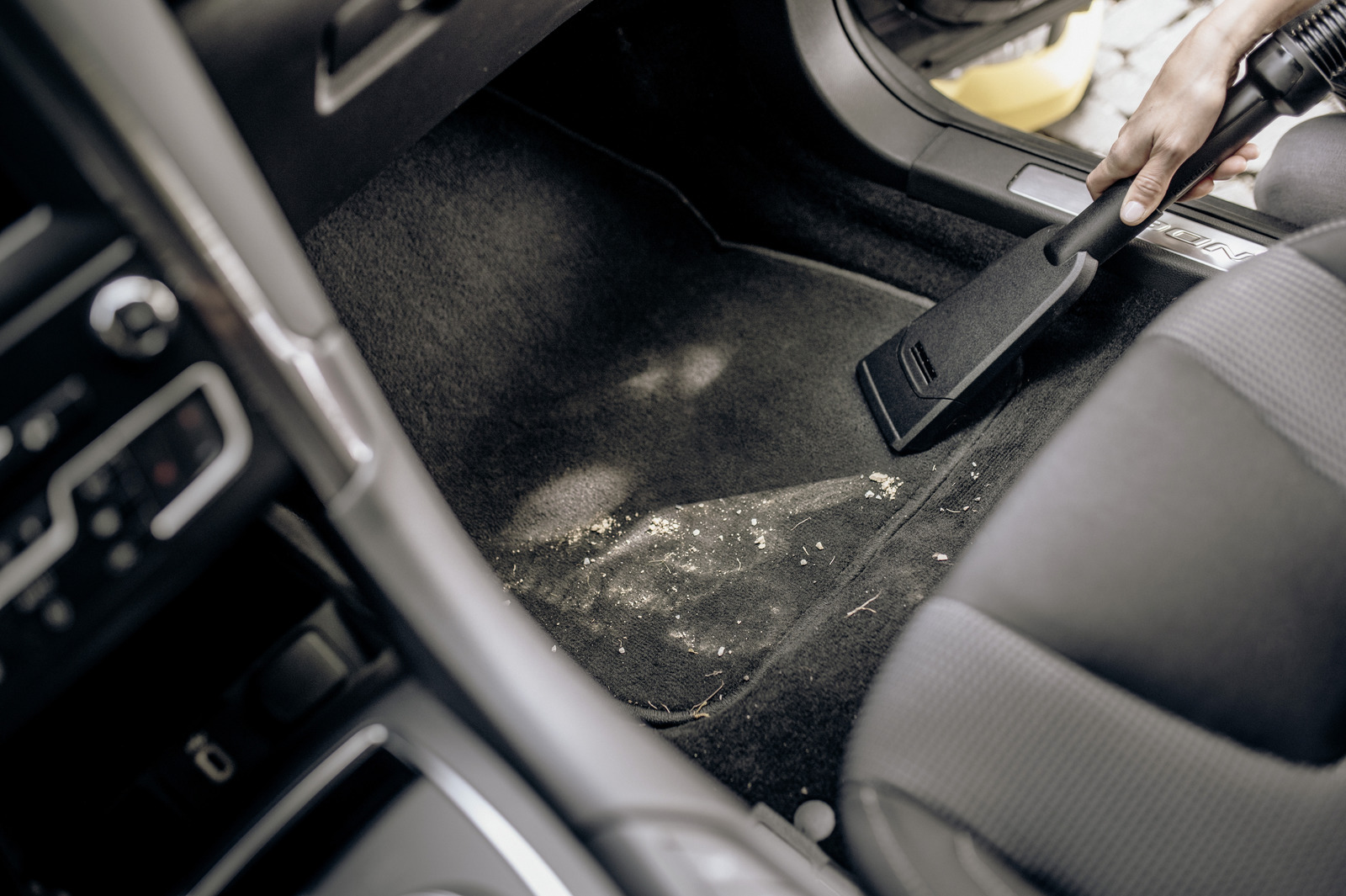 Osoba odkurza brudną, czarną matę podłogową w samochodzie. Na macie jest piasek i zanieczyszczenia. Siedzenie jest szare.