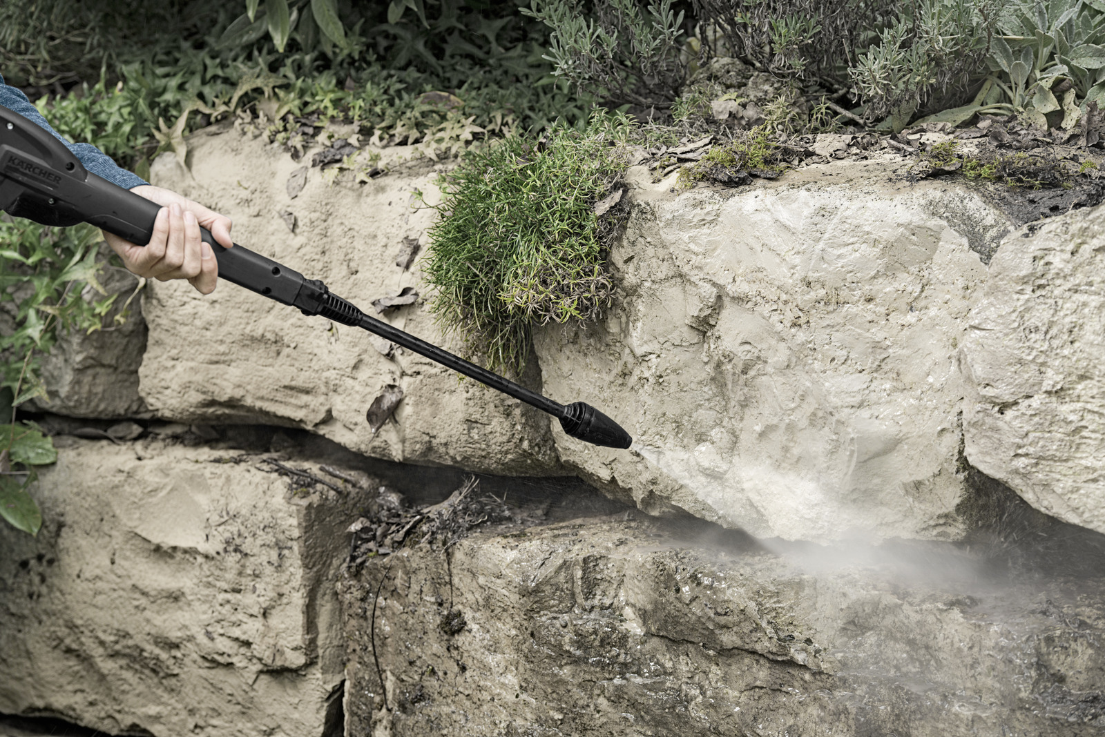 A person uses a pressure washer to clean a stone wall. The black nozzle sprays water, creating mist. Green plants and foliage are visible.