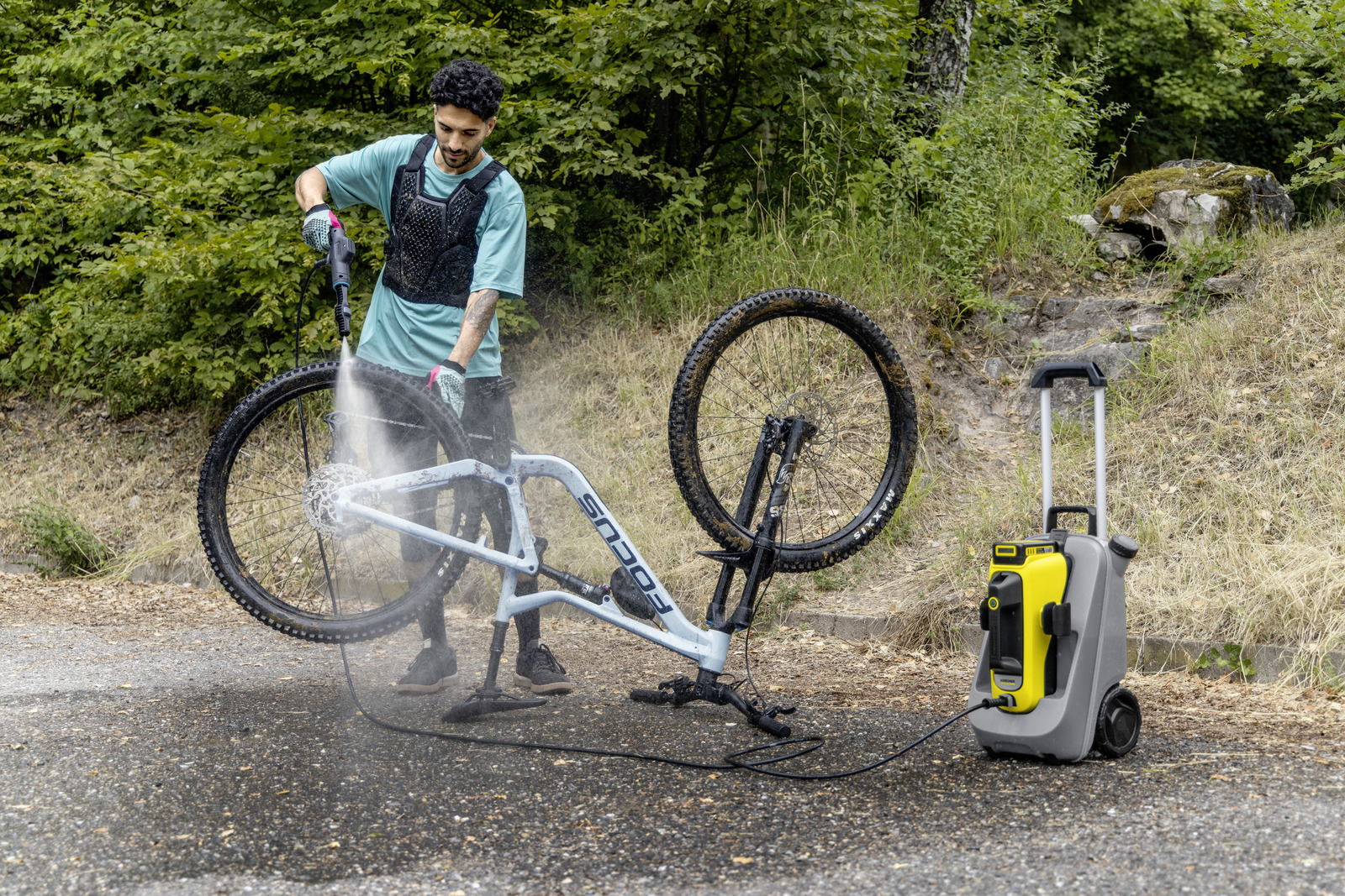 Ein Mann wäscht ein hellblaues Mountainbike mit einem Hochdruckreiniger. Eine gelb-graue Maschine steht neben dem Fahrrad auf nassem Untergrund.