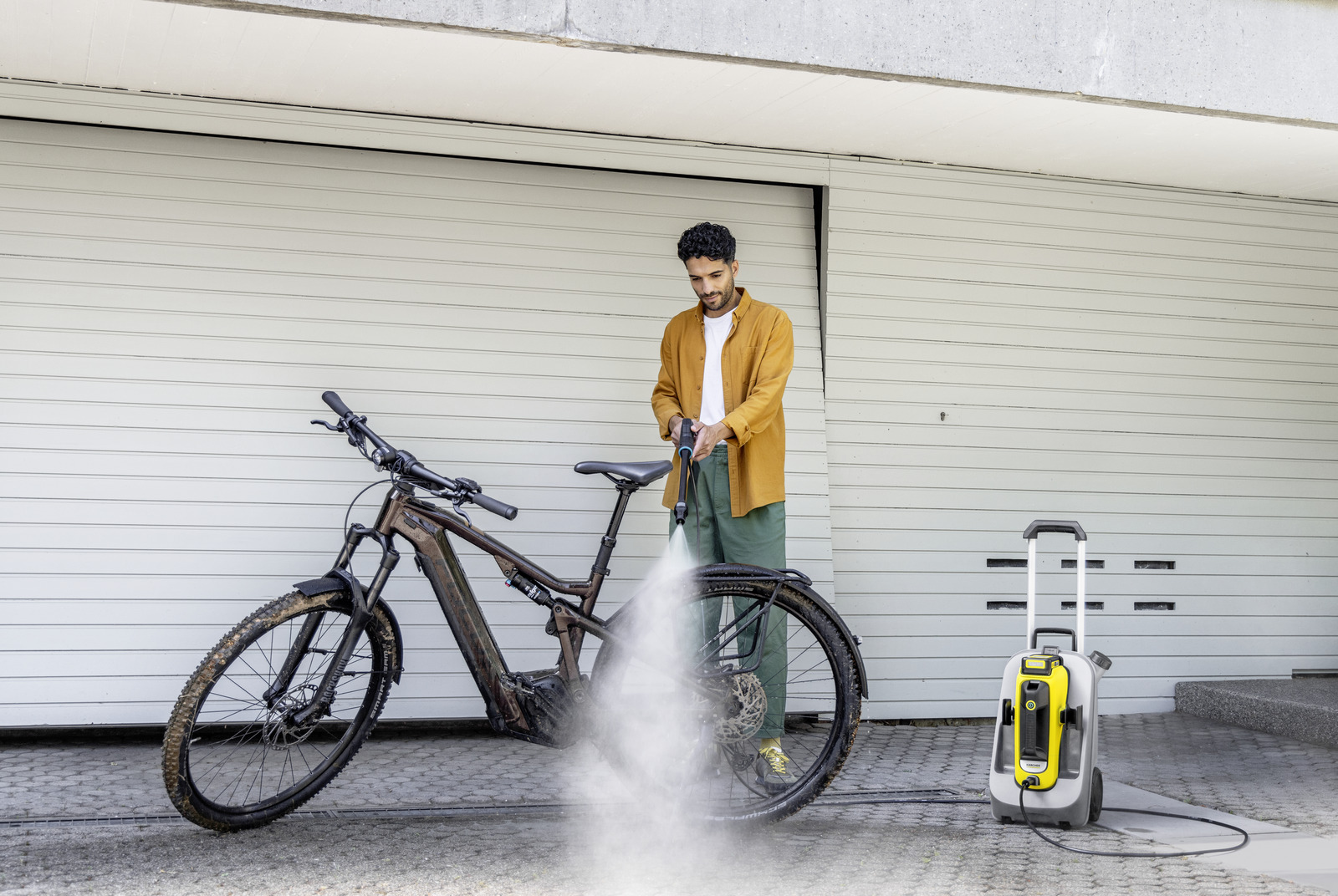 Ein Mann wäscht ein schlammiges Fahrrad mit einem Hochdruckreiniger. Rechts steht eine gelb-graue Maschine. Im Hintergrund ist ein Garagentor.
