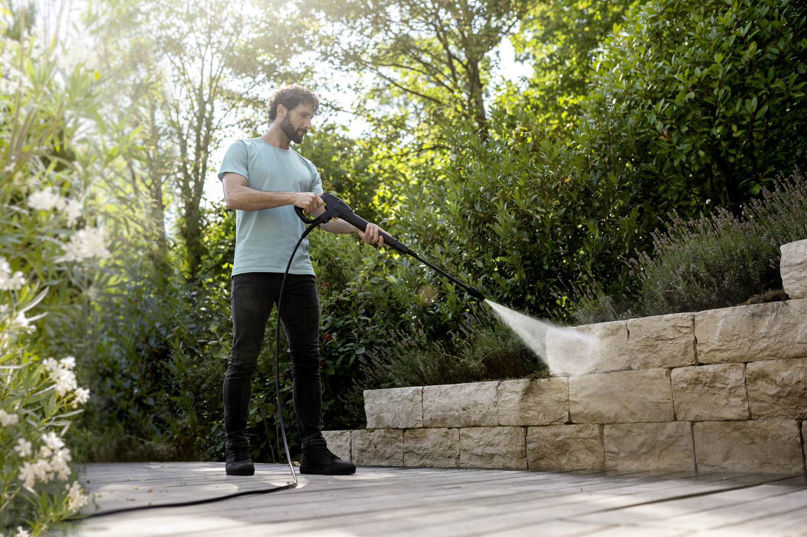 Mężczyzna używa myjki ciśnieniowej do czyszczenia kamiennego muru. Scena rozgrywa się na zewnątrz, z zielenią w tle i drewnianą podłogą.