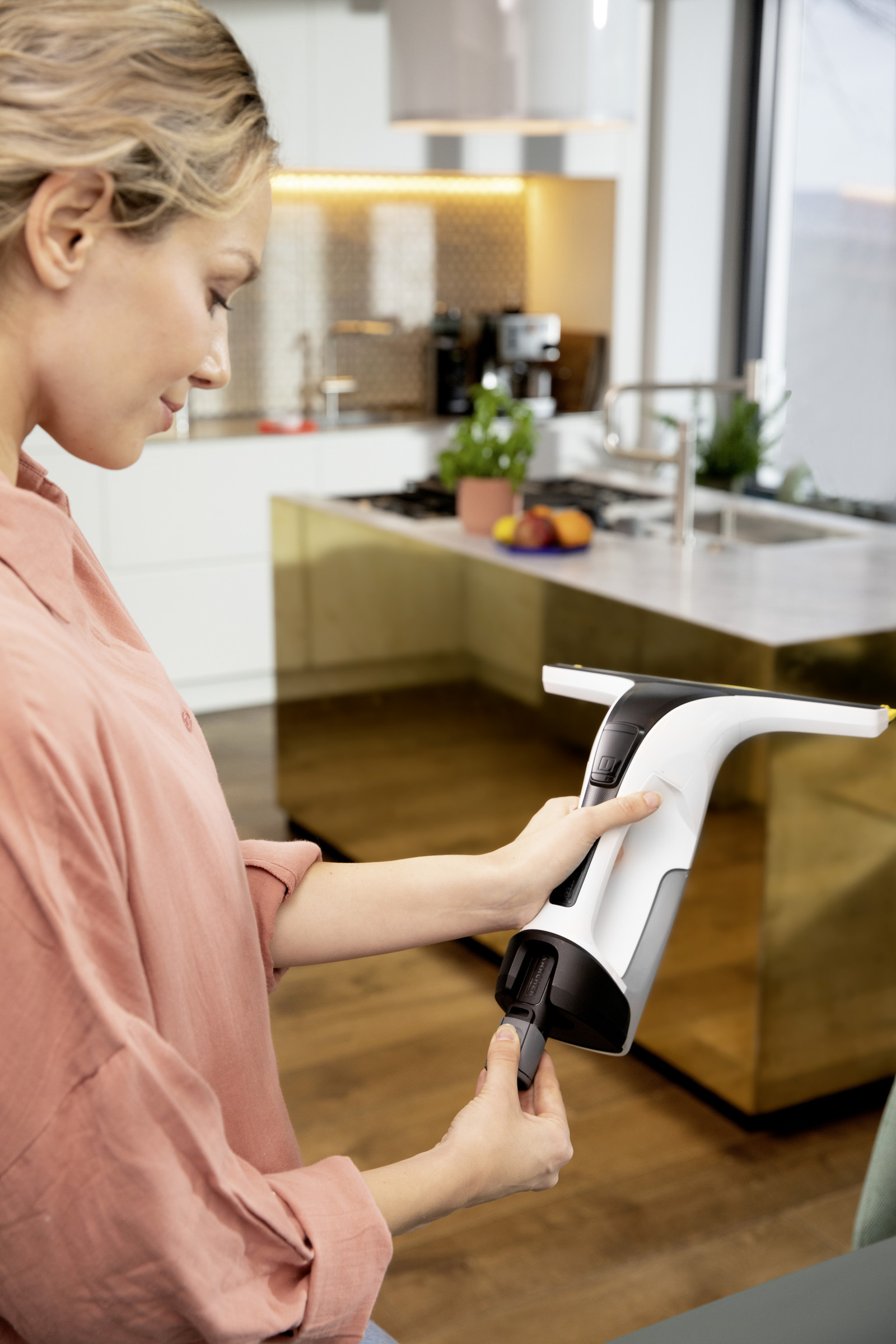 Kobieta trzyma biało-czarny środek do czyszczenia okien. Jest w nowoczesnej kuchni. Ma na sobie różową koszulę.