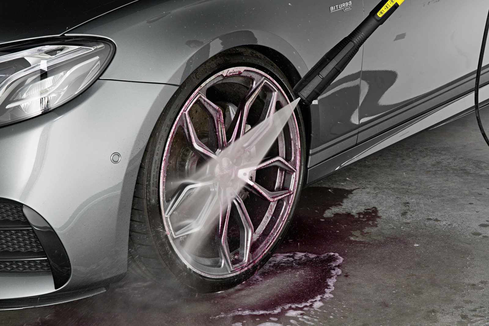 Ein graues Autorad wird mit einem Hochdruckreiniger gereinigt. Wasser sprüht auf das Rad und erzeugt einen Nebel. Rosa Flüssigkeit läuft auf dem Boden.