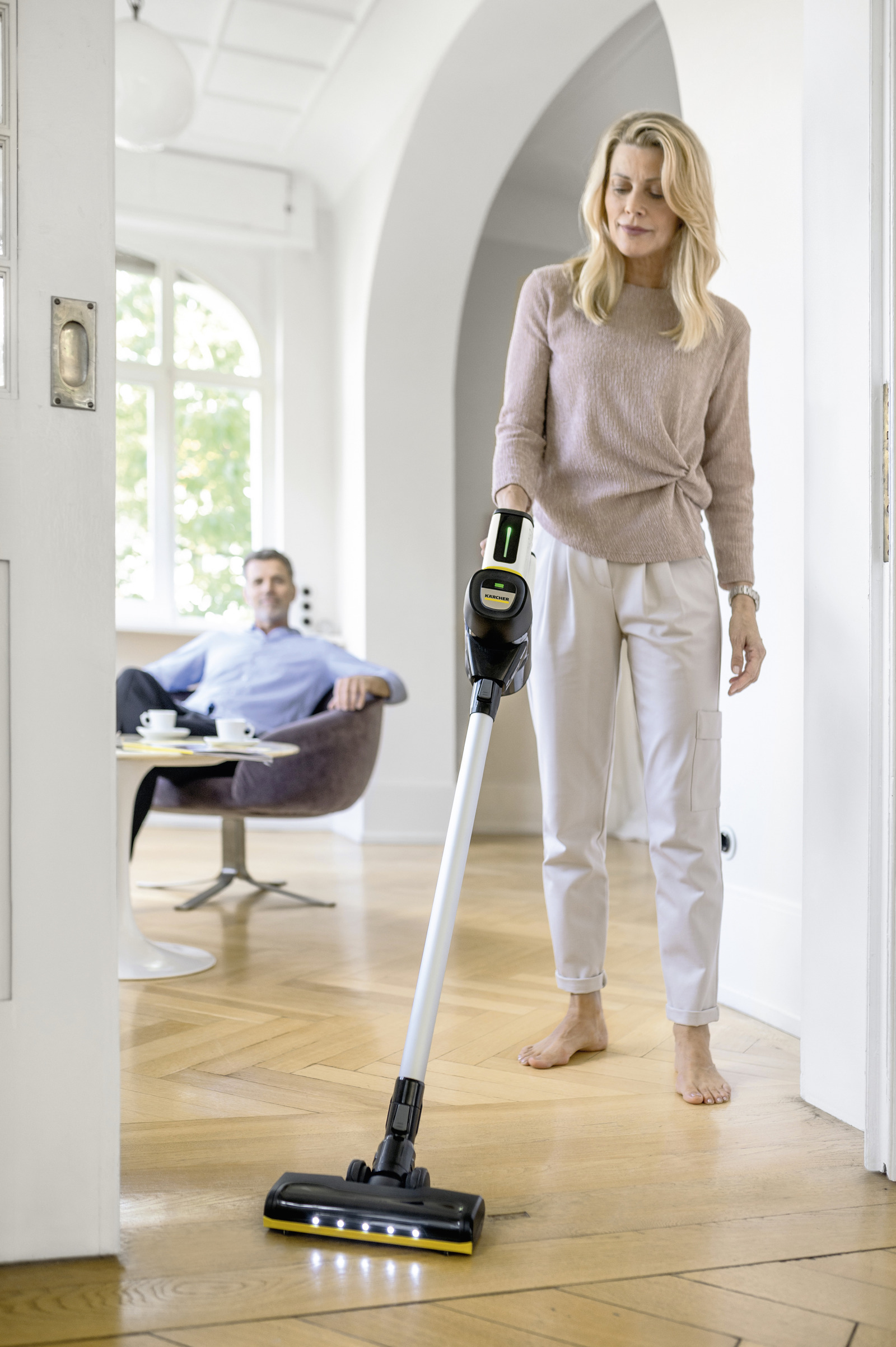 Une femme passe l'aspirateur sur un sol en bois. Un homme est assis en arrière-plan. L'aspirateur est noir et jaune.
