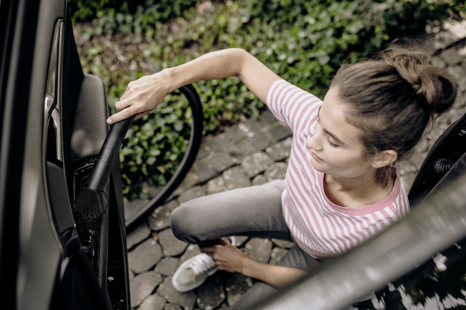 Een vrouw stofzuigt het interieur van een auto. Ze draagt een gestreept shirt en een grijze broek. De achtergrond toont groen en stenen bestrating.