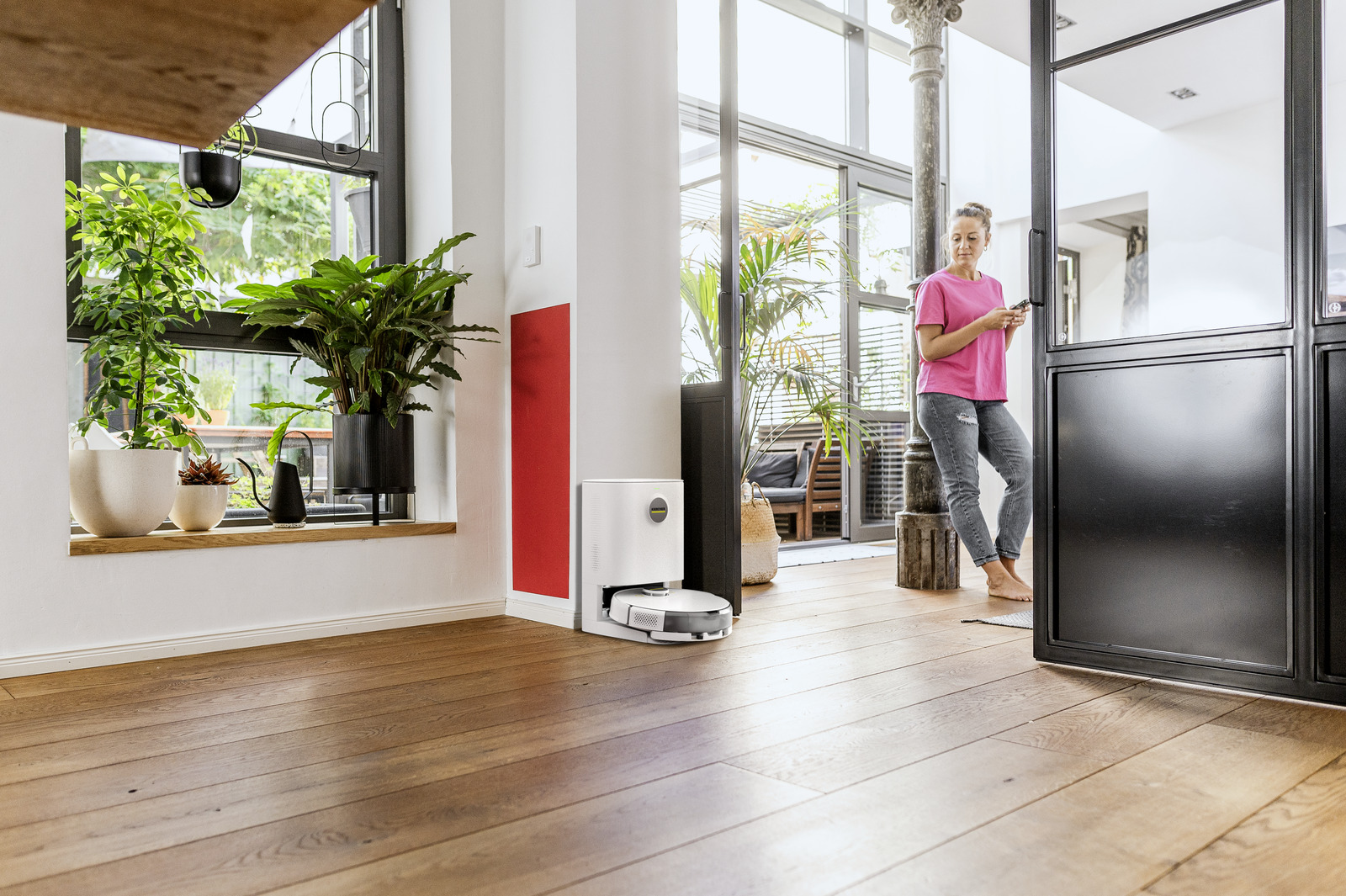 Una donna con top rosa e jeans è appoggiata a un telaio nero, tiene un telefono. Un robot aspirapolvere è sul pavimento.