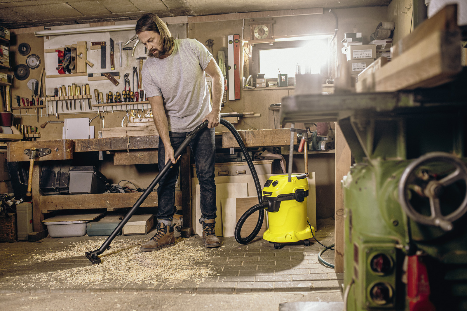 Un uomo aspira il pavimento di un'officina coperto di trucioli di legno. Un aspirapolvere giallo è vicino a lui. Attrezzi appesi.