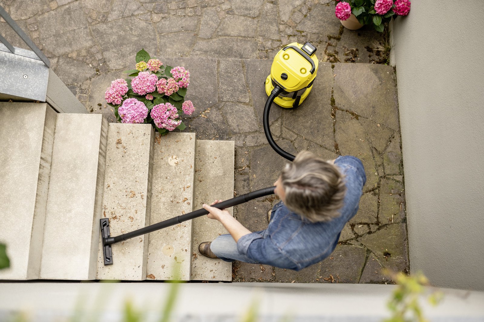 Una persona aspira escalones al aire libre. Una aspiradora amarilla está en un escalón. Cerca hay flores rosas en macetas.