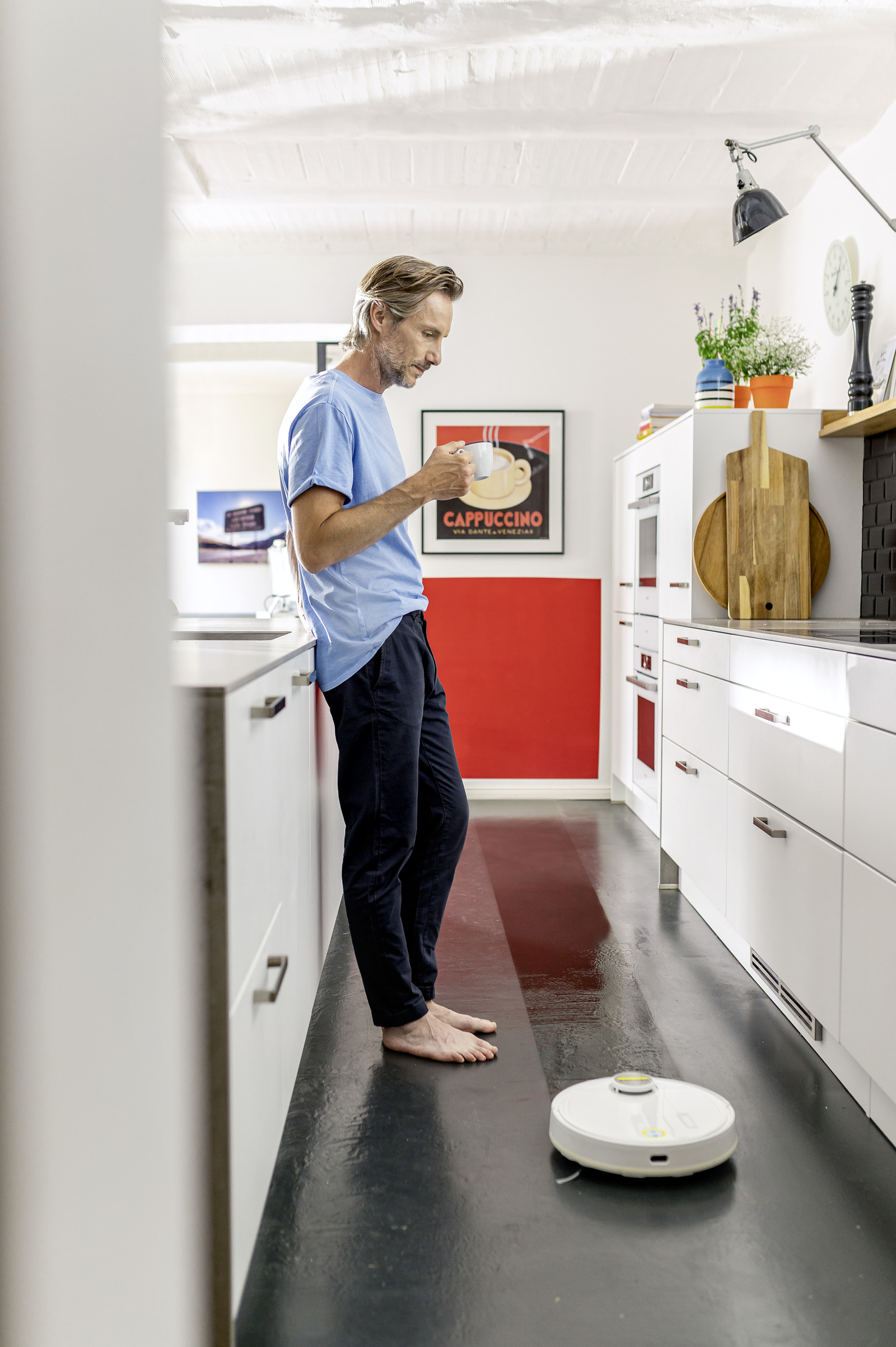 Hombre con camisa azul se apoya en la encimera de la cocina, sostiene una taza. Un robot aspirador limpia el suelo. Cartel de capuchino.