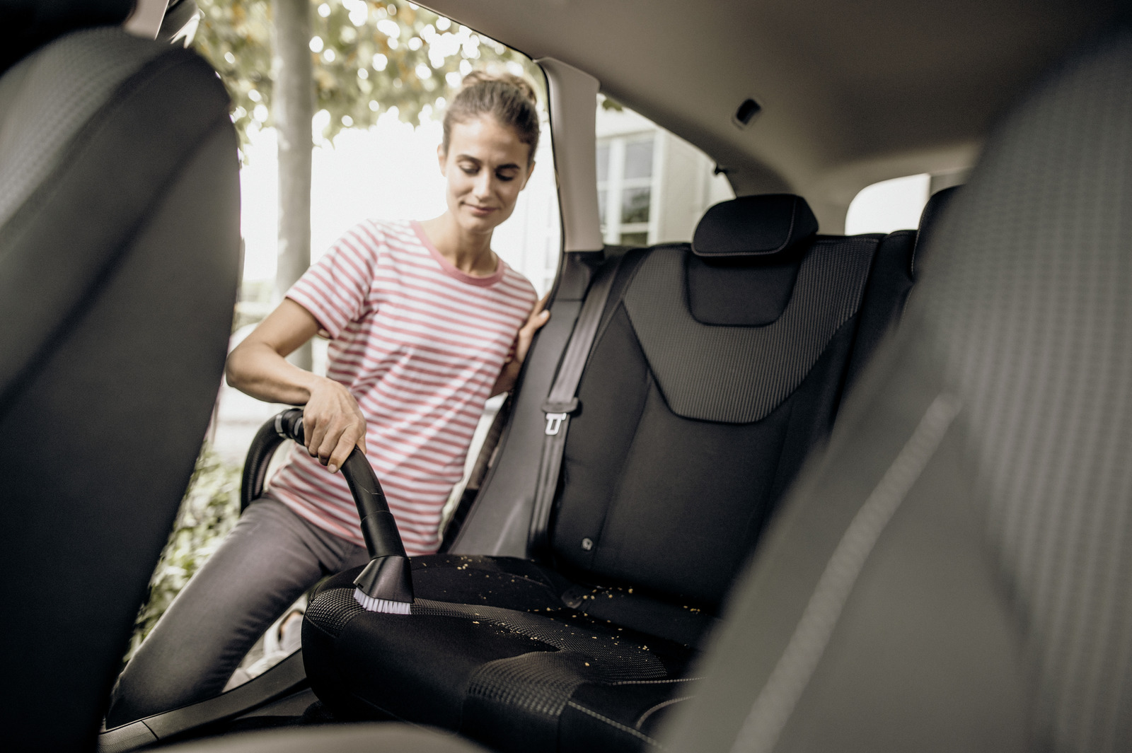 Una mujer aspira un asiento de coche. Lleva una camisa a rayas. El asiento es negro y el interior del coche es visible.