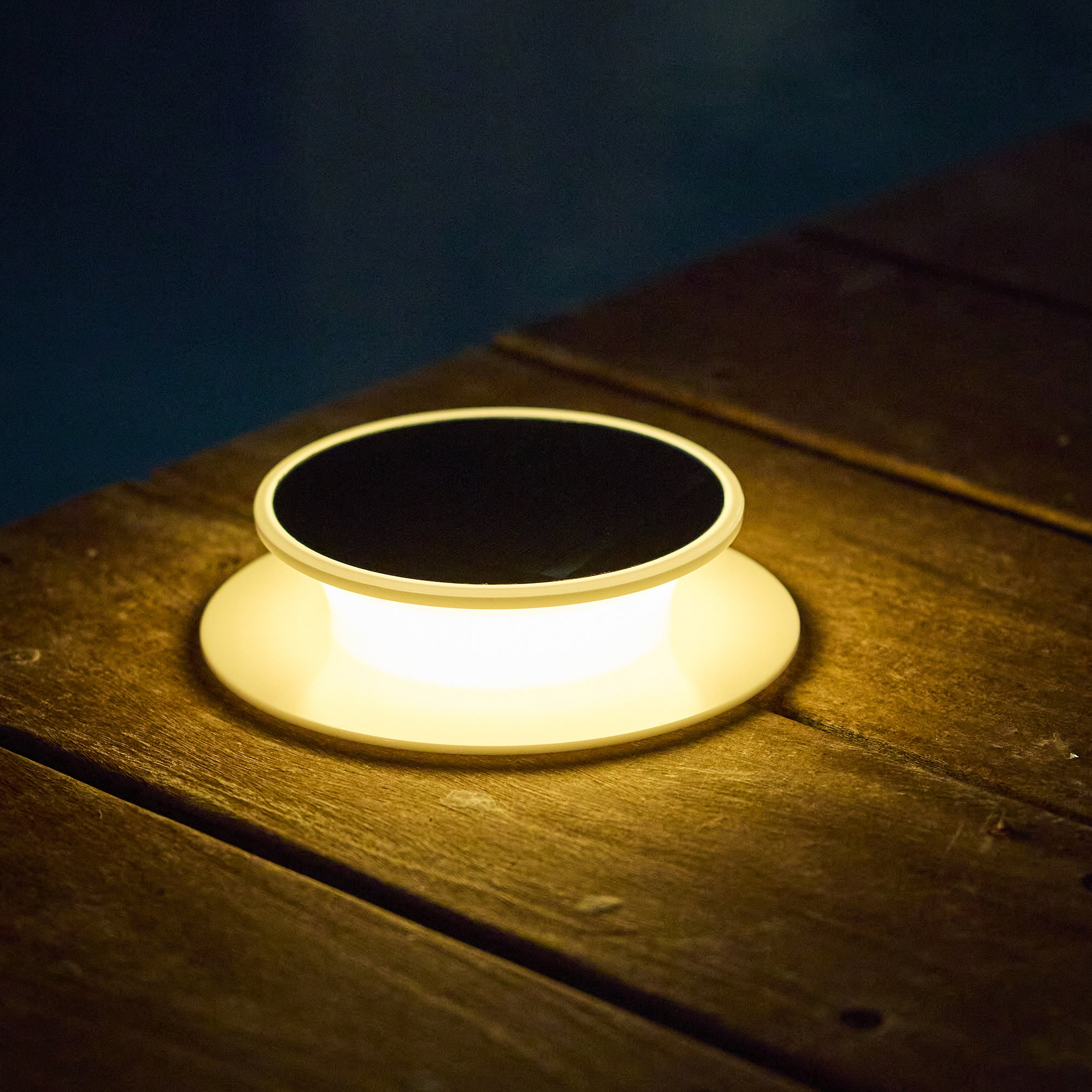 Eine leuchtende Lampe mit schwarzem Deckel steht auf Holzplanken. Der Hintergrund ist dunkelblau, möglicherweise Wasser.