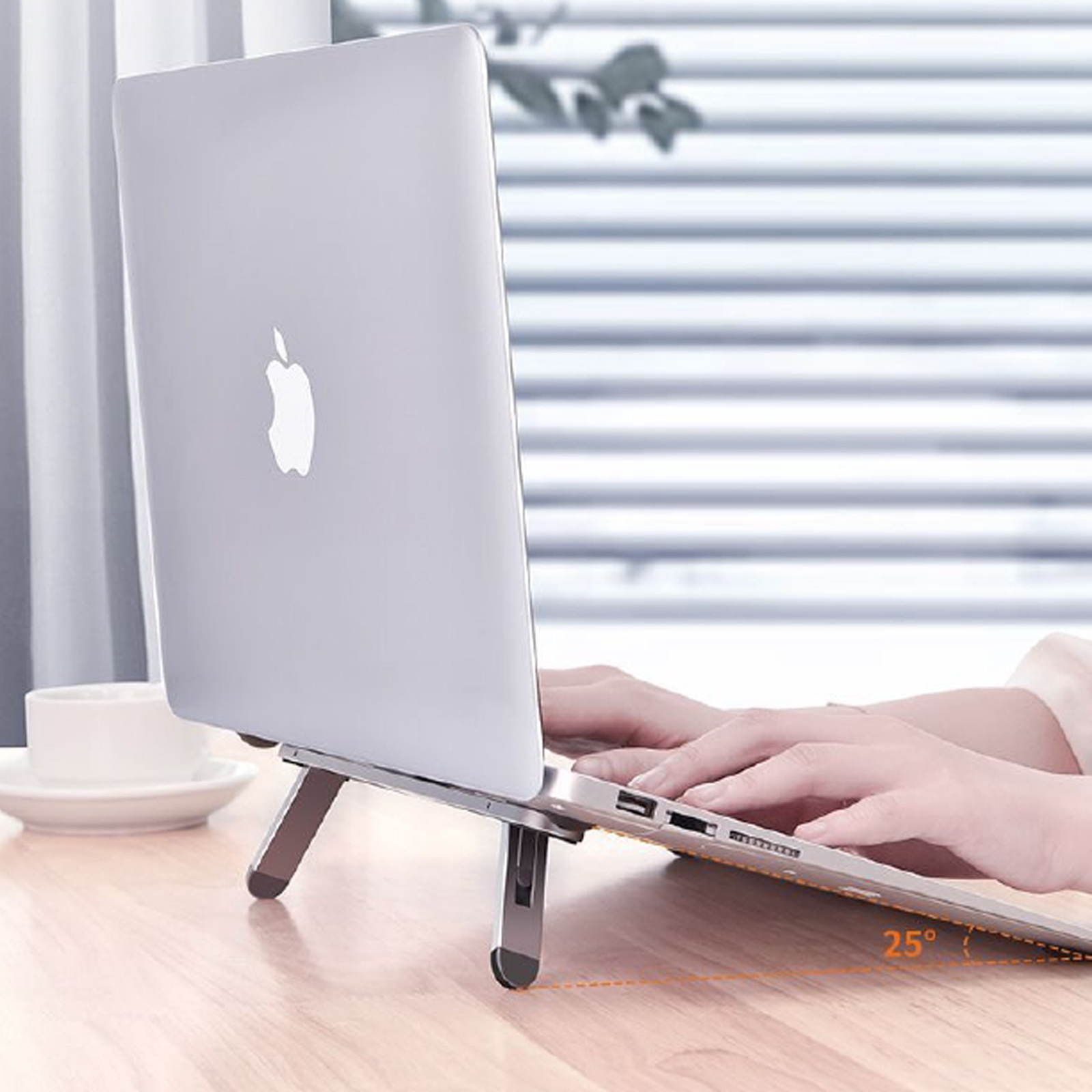 Una laptop sobre un soporte, inclinada 25 grados. Manos escribiendo. Una taza y platillo a la izquierda.