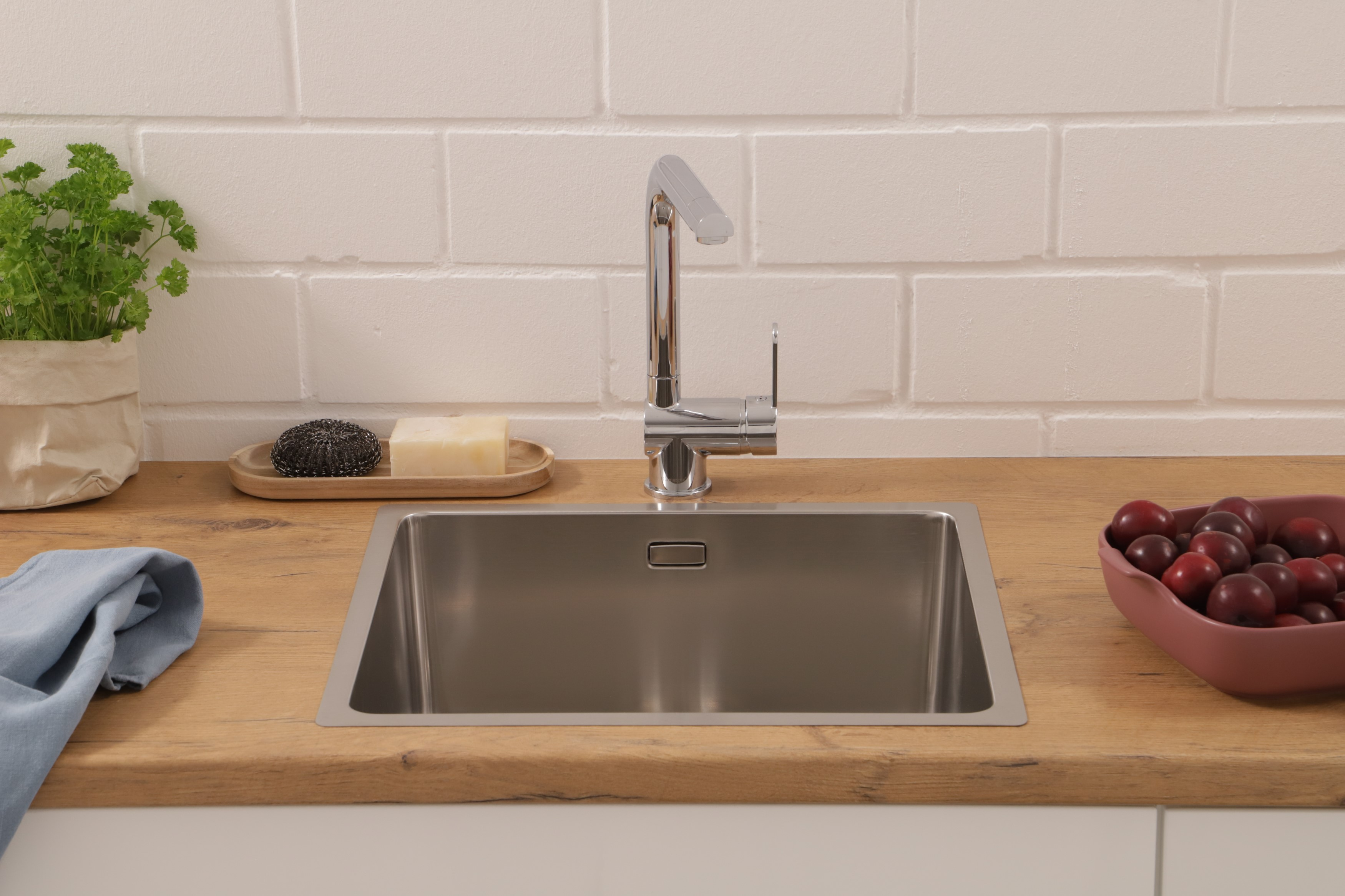 Ein Küchenbecken mit Wasserhahn, Seife, einer Pflanze und einer Schüssel Pflaumen auf einer Holzplatte. Weiße Backsteinmauer.
