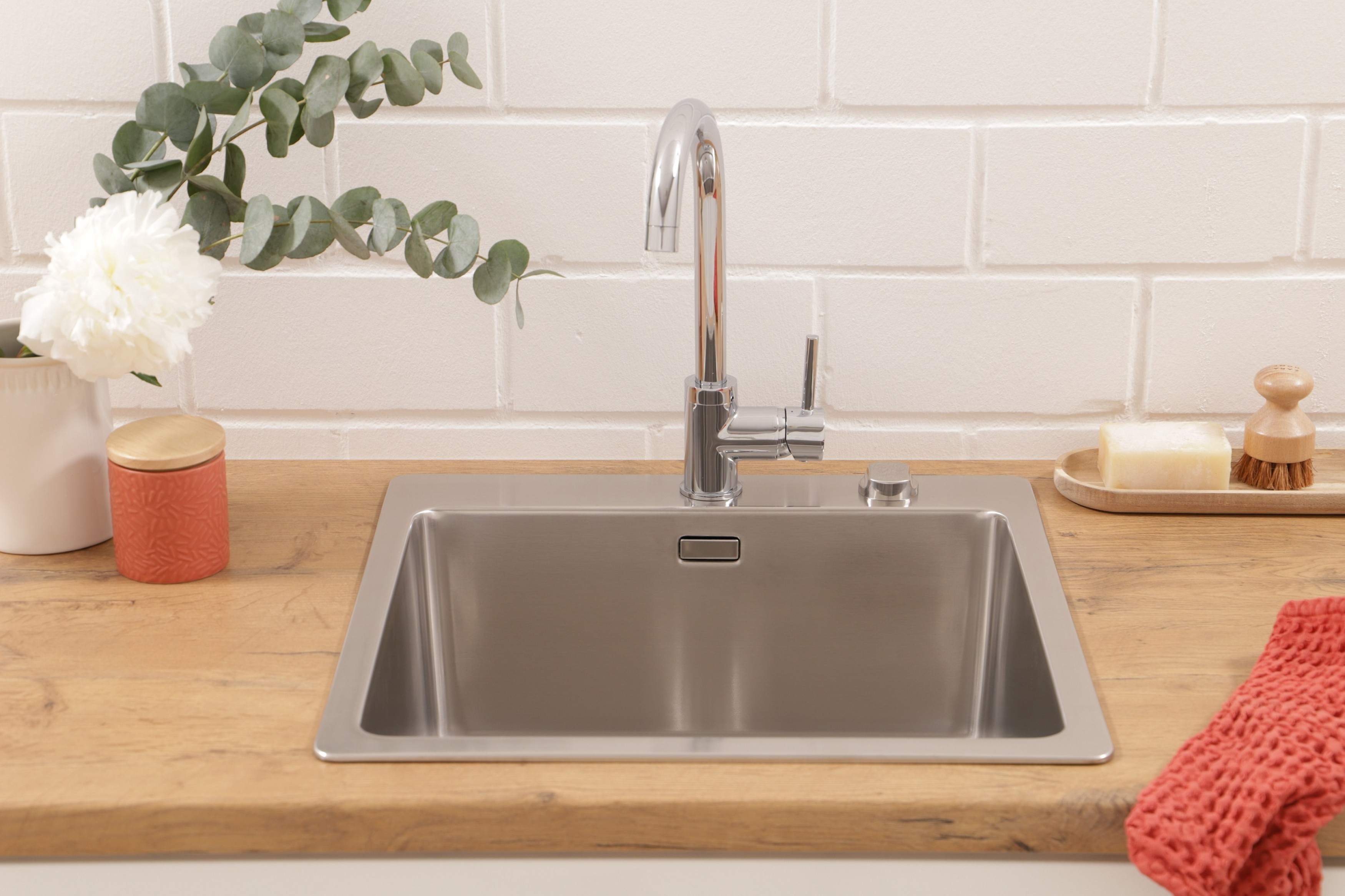 Ein Küchenbecken mit Wasserhahn, Seife, Bürste und einem roten Tuch auf einer Holztheke mit Blumen.