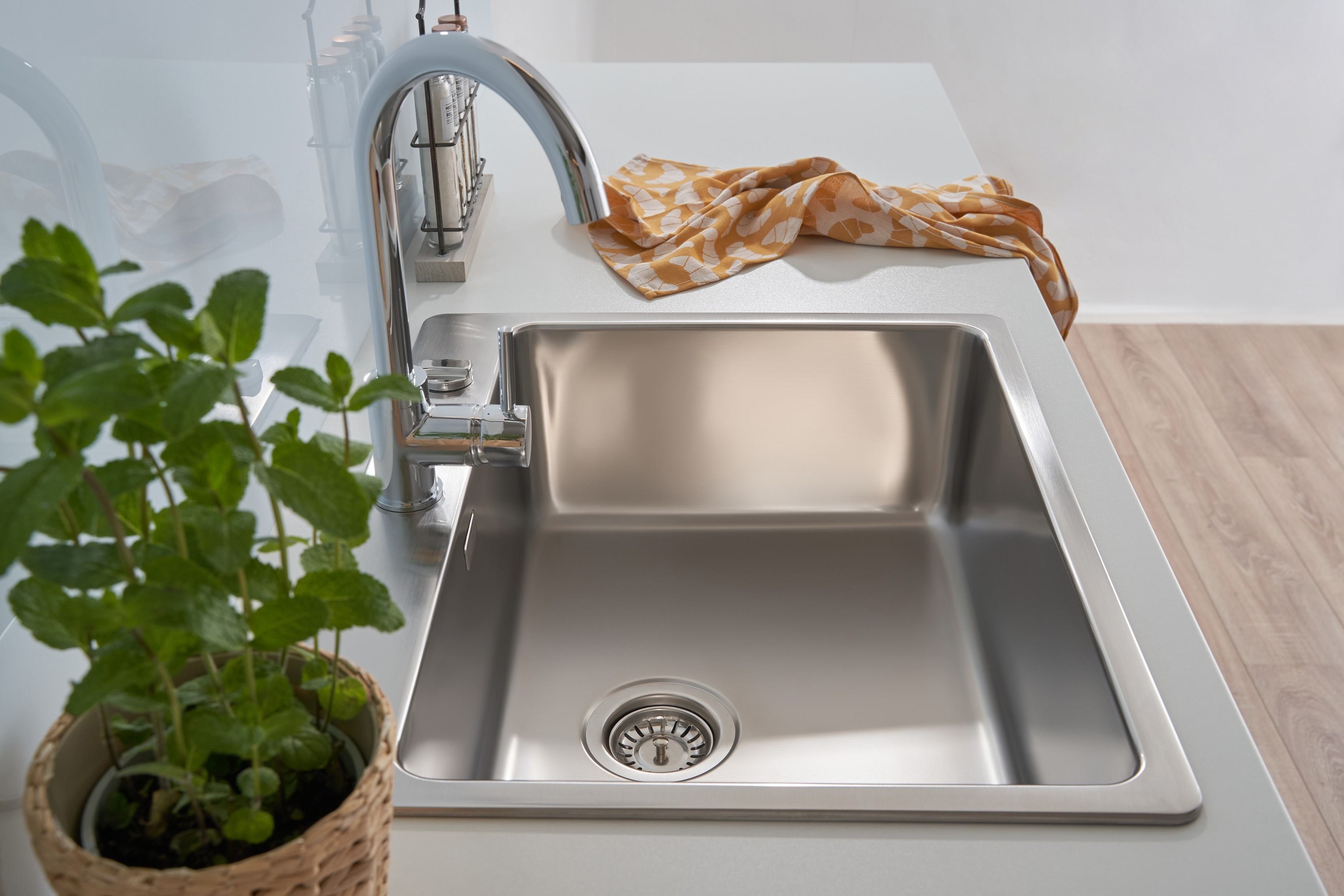 Ein Edelstahlbecken mit Wasserhahn, gelb gemustertem Tuch und Topfpflanze auf weißer Theke.