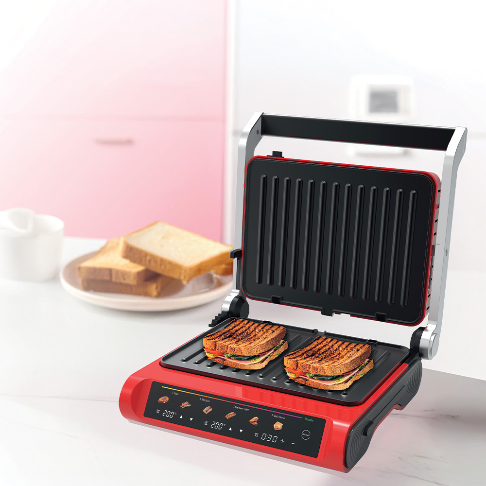 İçinde iki sandviç bulunan kırmızı bir panini presi. Arka planda bir tabakta dilimlenmiş ekmek, pembe dolap.