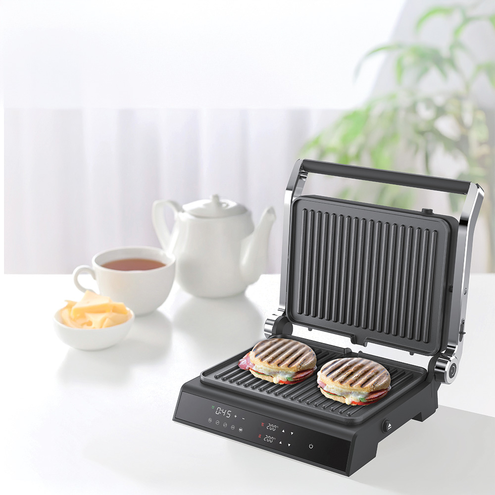 Beyaz bir yüzeyde iki sandviç, çay, peynir ve bir demlikle panini ızgarası. Arka planda yeşil bitki ve beyaz perdeler.