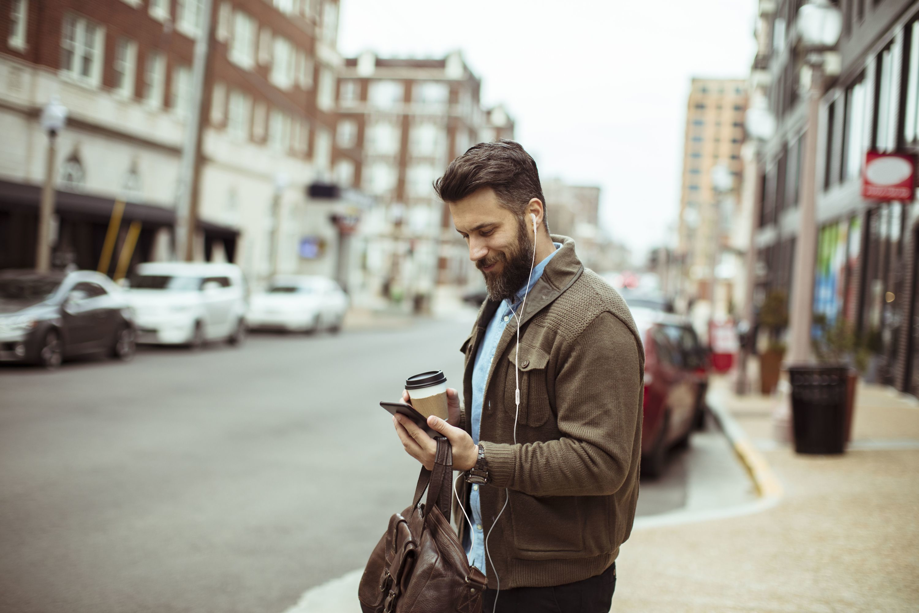 Mężczyzna z brodą, trzymający telefon i kawę, idzie ulicą. Budynki i samochody w tle.