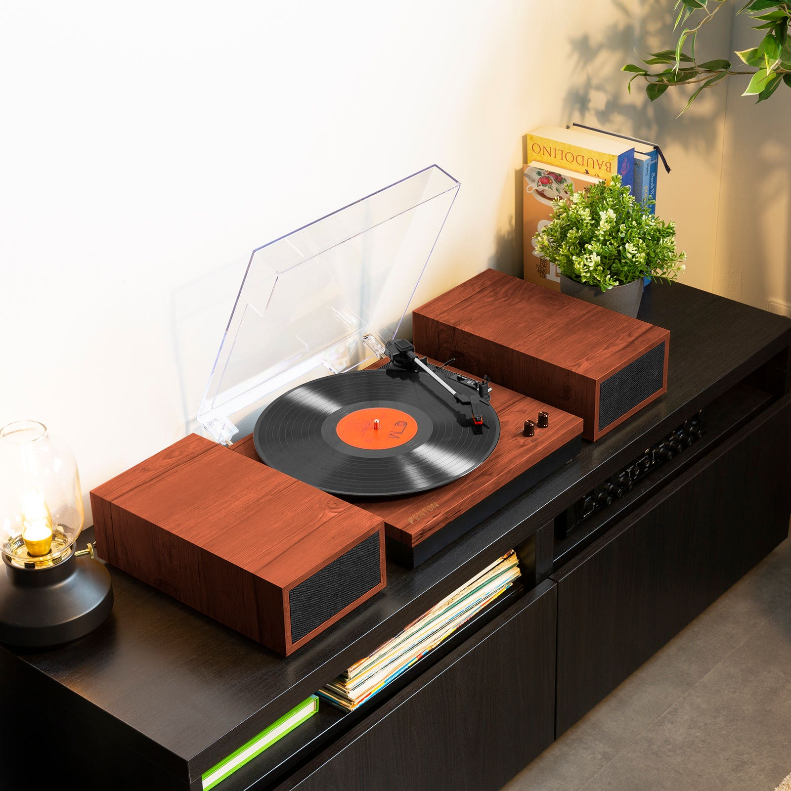 A turntable with a record playing, speakers, books, and a lamp on a black cabinet. Plant in background.
