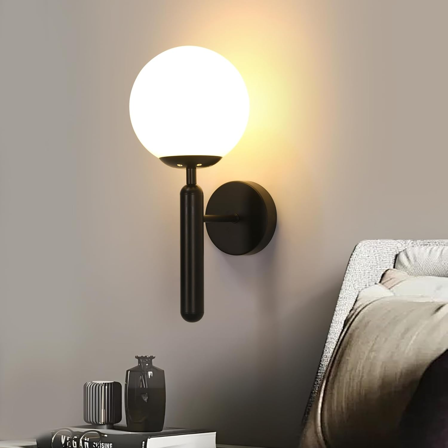 Eine moderne Wandlampe mit rundem, weißem Glasschirm. Sie ist an einer schwarzen Metallhalterung befestigt. Daneben befindet sich ein Bett und Dekoration.
