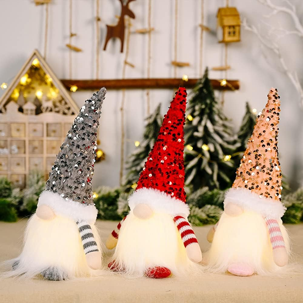 Drei beleuchtete Wichtel mit Paillettenhüten und flauschigen Bärten. Ein Hut ist grau, einer rot, einer rosa. Der Hintergrund hat Weihnachtsdekorationen.