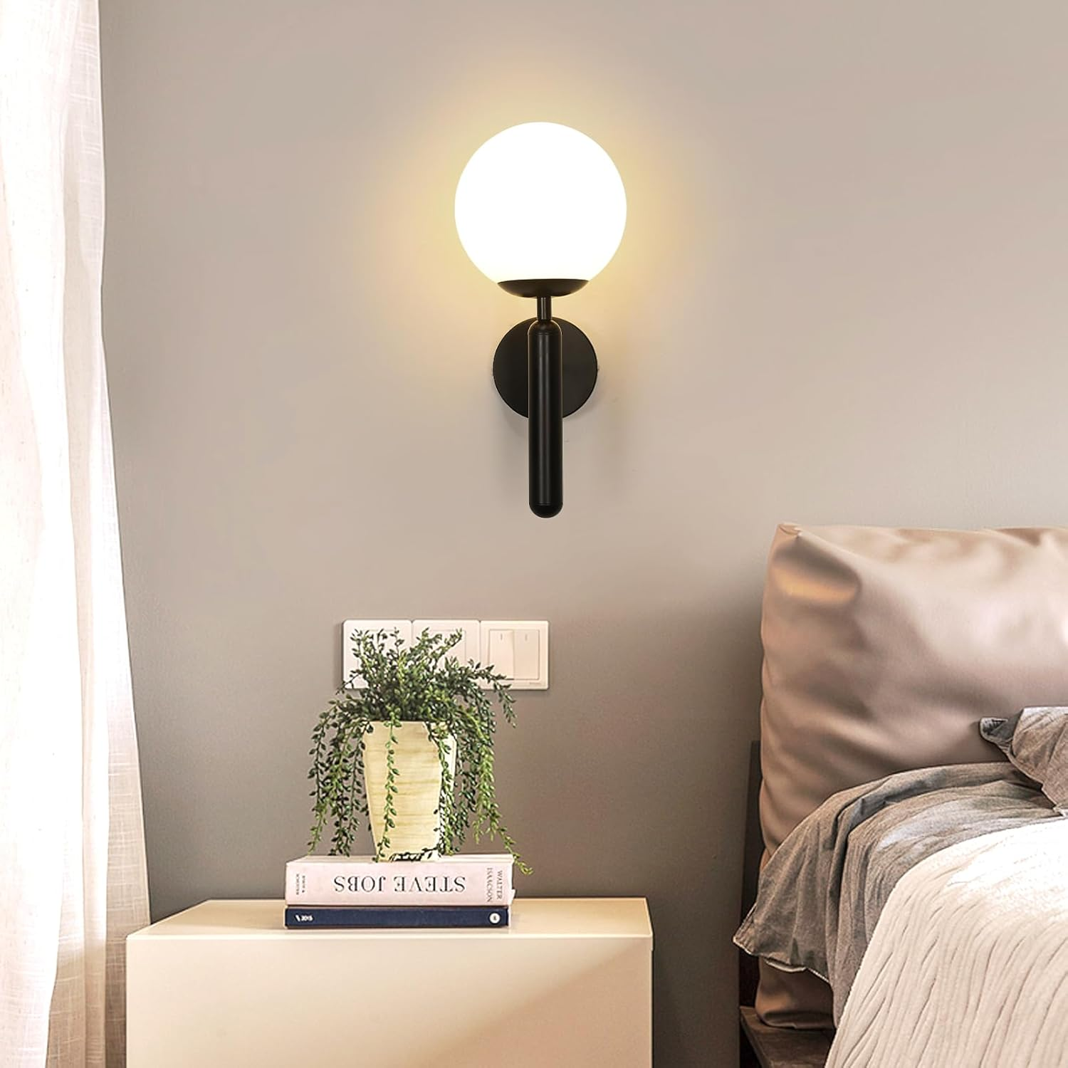 Ein modernes Schlafzimmer mit einer Wandleuchte, einem Nachttisch mit Büchern und einer Pflanze sowie einem Bett mit Kissen und Decken.