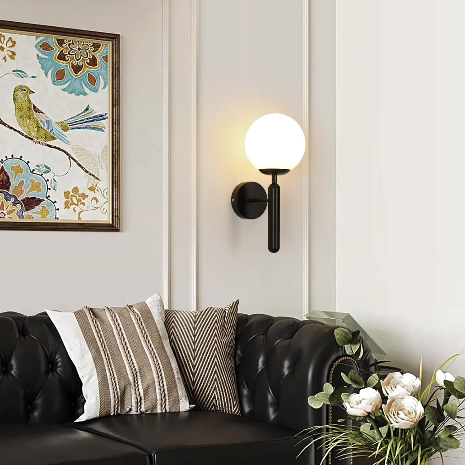Ein Raum mit schwarzem Ledersofa, Kissen, Wandlampe, Vogelgemälde und Blumen.