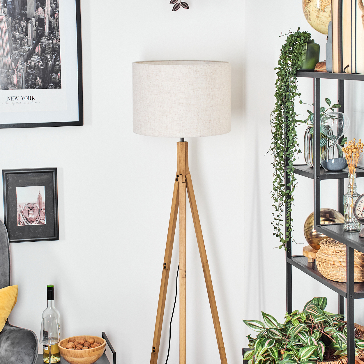 Ein Raum mit einer hölzernen Stativ-Stehlampe. Ein Regal mit Pflanzen, Vasen und Dekorationsartikeln. Wandkunst und eine Schüssel mit Walnüssen sind vorhanden.