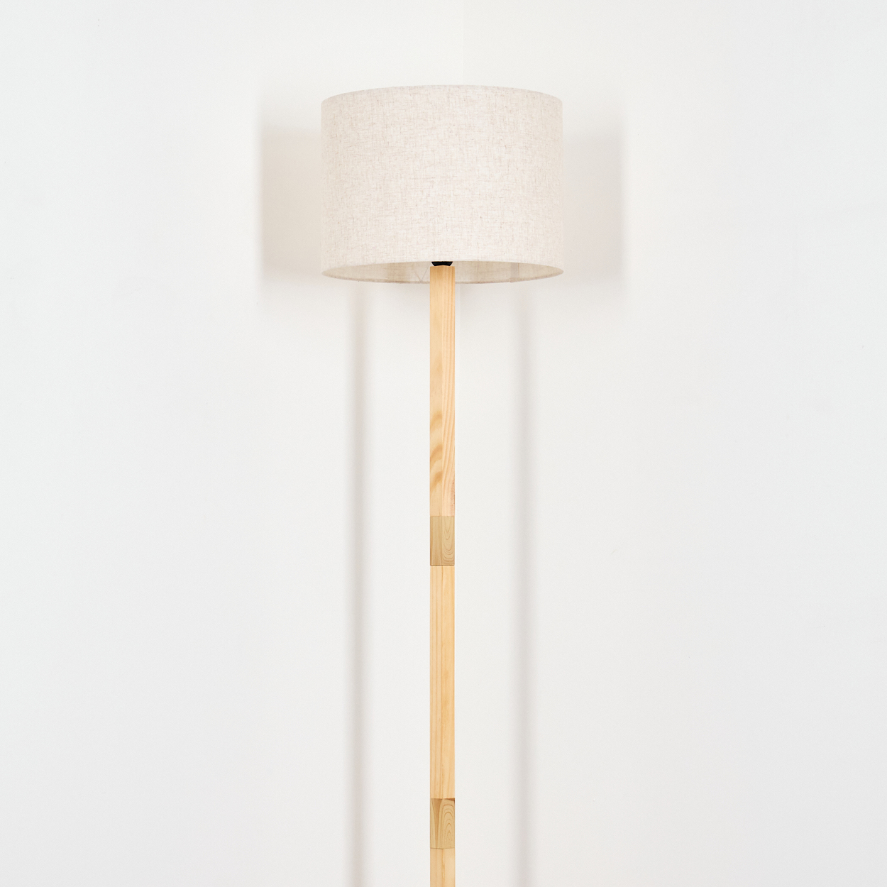 Ein beigefarbener Lampenschirm sitzt auf einem hölzernen Lampenständer vor einer schlichten weißen Wand. Die Lampe ist zentriert und beleuchtet.