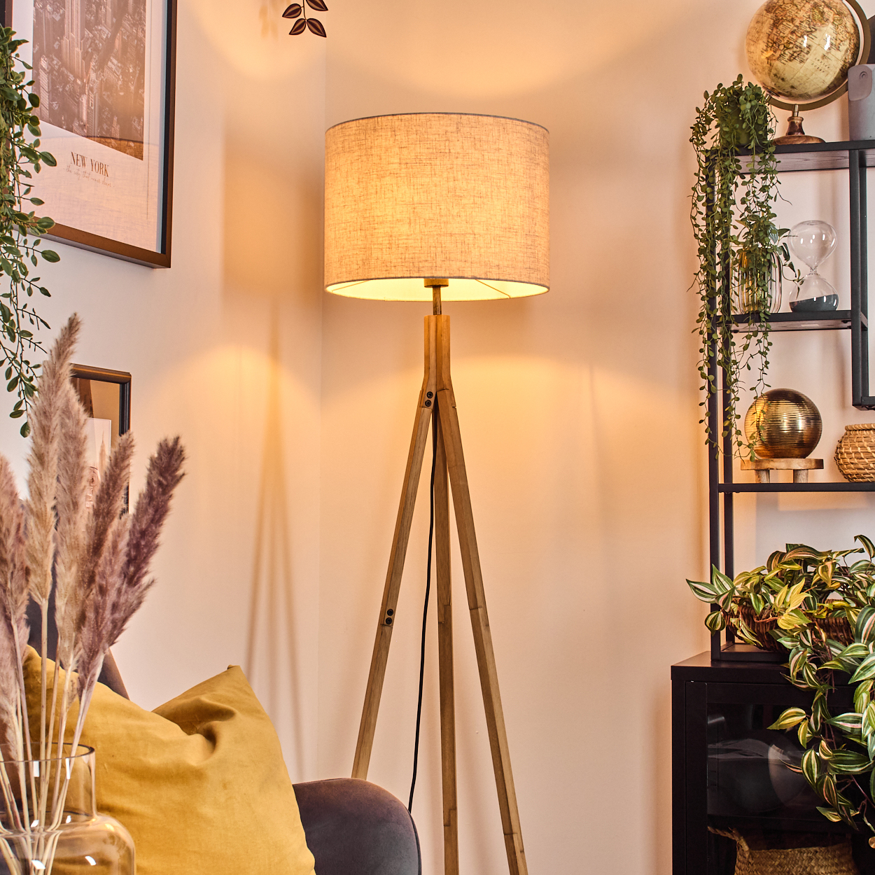 Eine Zimmerecke mit einer hölzernen Stativlampe, Pflanzen, einem Globus und gerahmten Bildern. Ein gelbes Kissen und Pampasgras sind sichtbar.