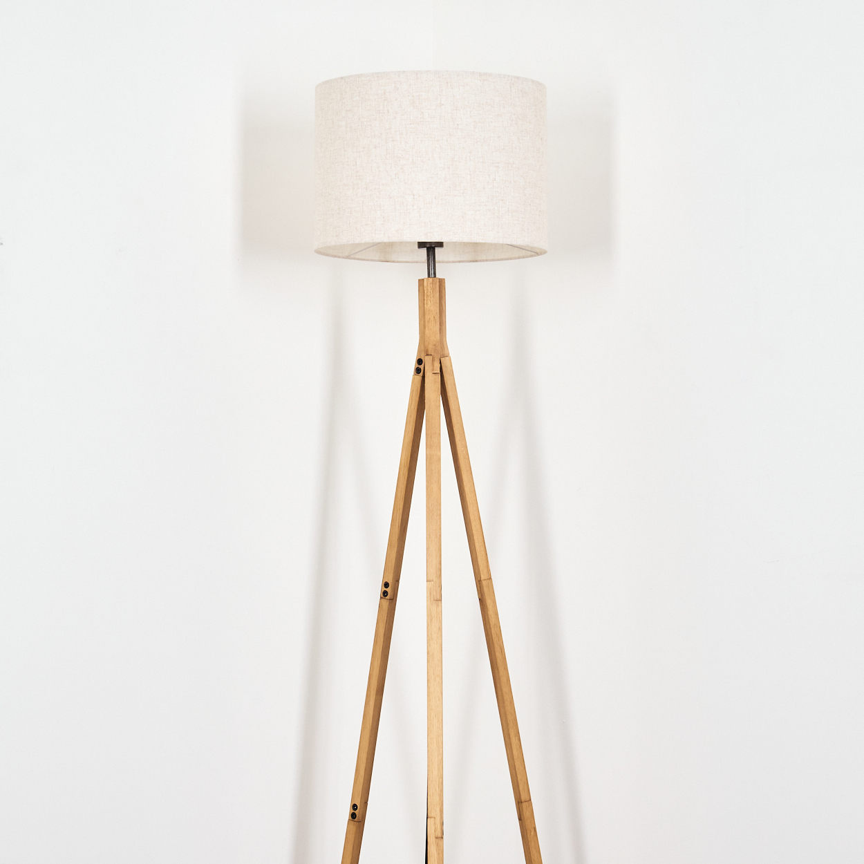 Eine hölzerne Stativ-Stehlampe mit beigem Lampenschirm steht vor einer weißen Wand. Die Lampe hat drei Beine.