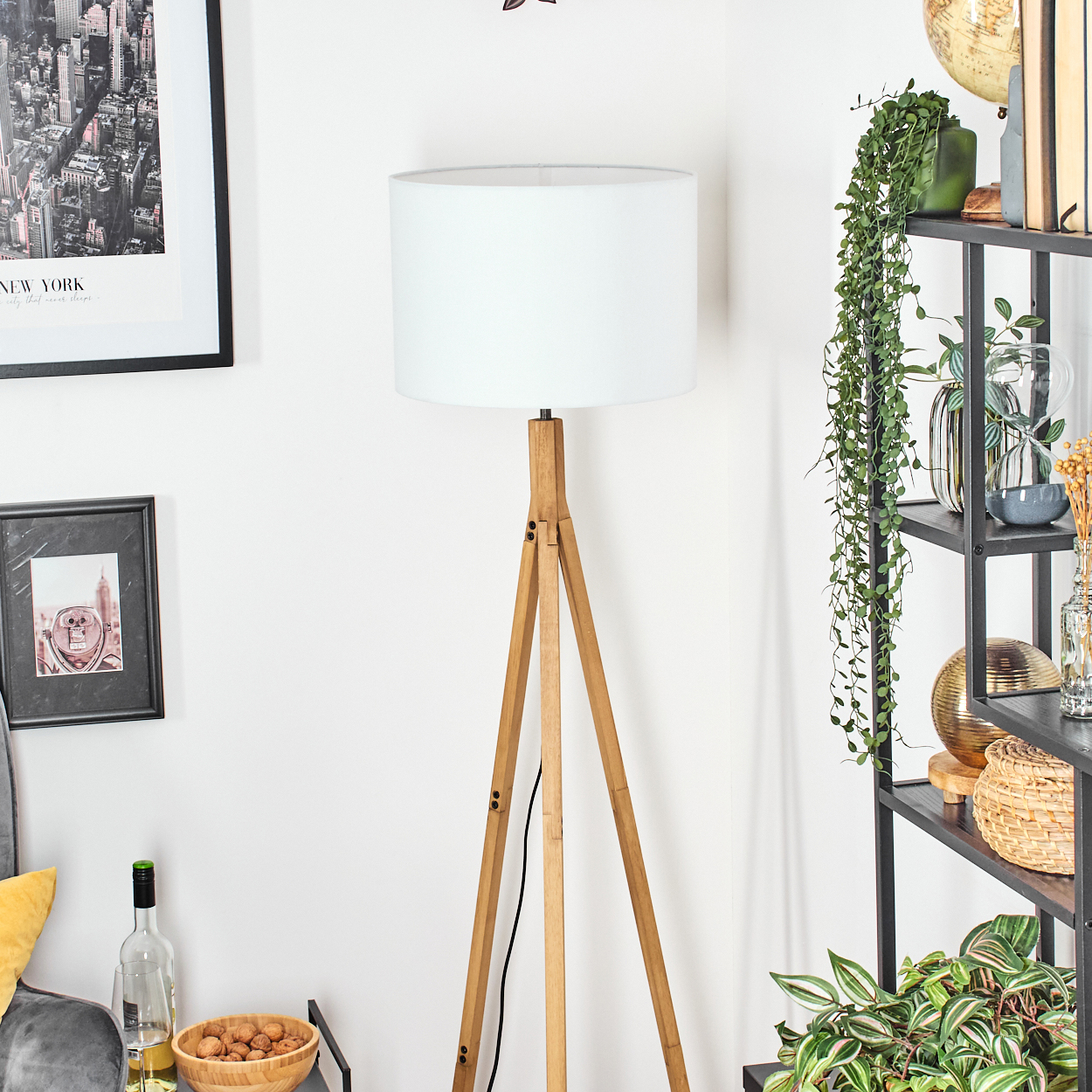Eine weiße Lampe mit Holzbeinen steht vor einer weißen Wand. Regale mit Pflanzen und Dekoration sind rechts sichtbar.