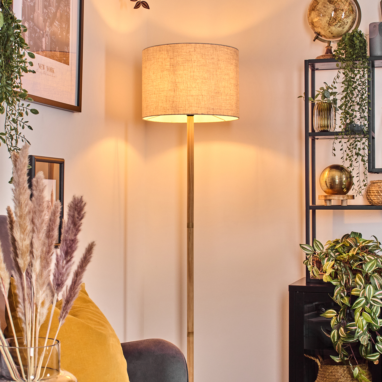 Eine Stehlampe steht in einem Raum mit Regal, Pflanzen und einem New York-Druck. Ein gelbes Kissen liegt auf einem grauen Stuhl.