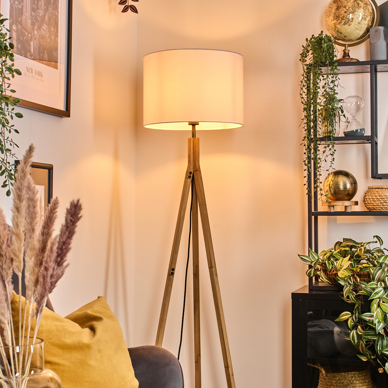 Eine beleuchtete Holzstativlampe steht in einer Ecke. Rechts befindet sich ein Regal mit Pflanzen und Dekoration. Links sind ein New York-Bild und Pampasgras.