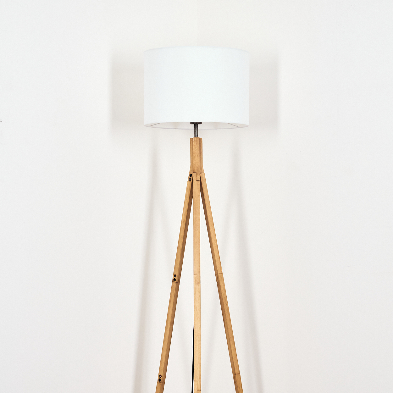 Eine Stehlampe aus Holz mit weißem Lampenschirm steht vor einer weißen Wand. Die Lampe hat ein schlichtes, modernes Design.