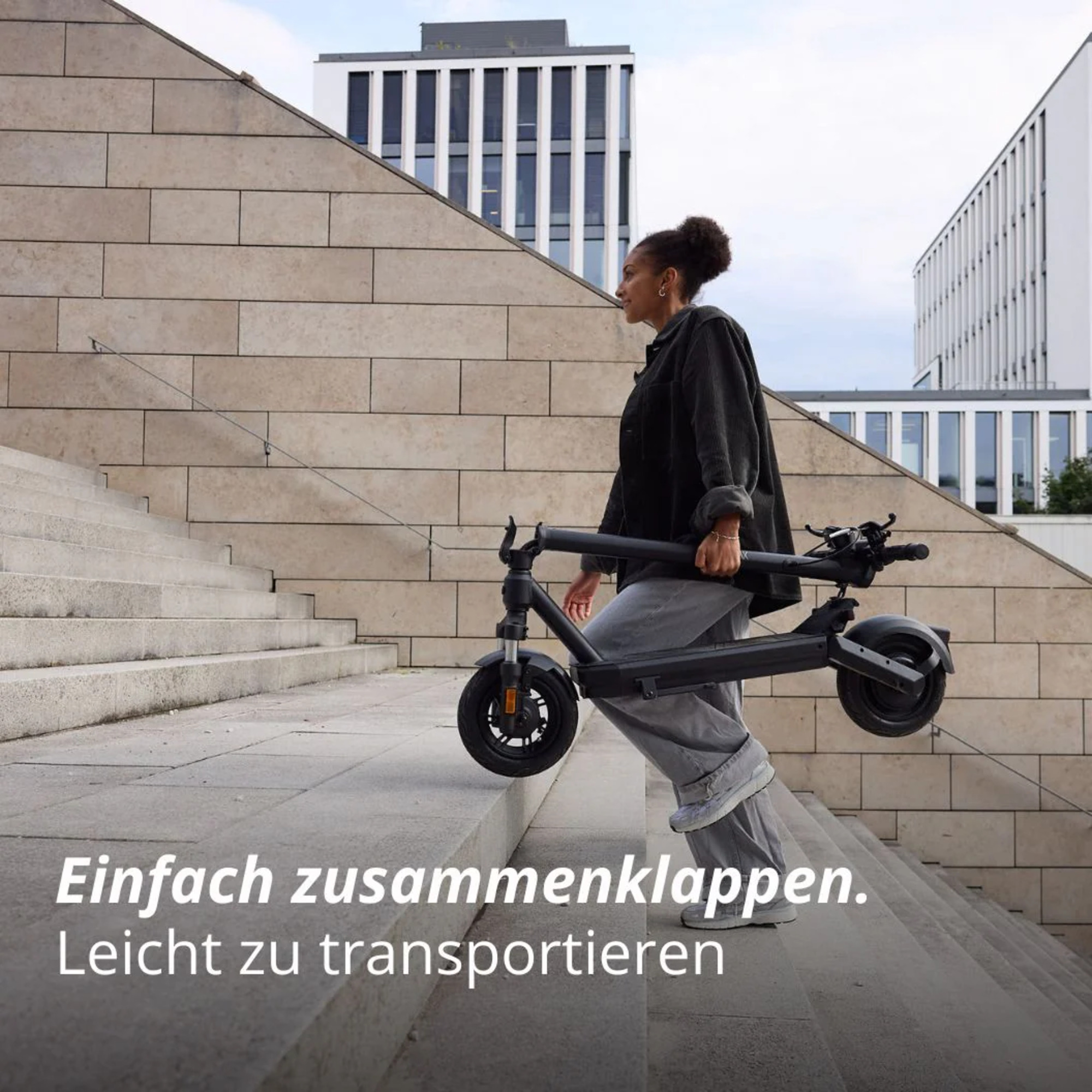 Eine Frau trägt einen zusammengeklappten schwarzen Roller die Treppe hinauf. Gebäude im Hintergrund. Text: Einfach zusammenklappen, leicht zu transportieren.
