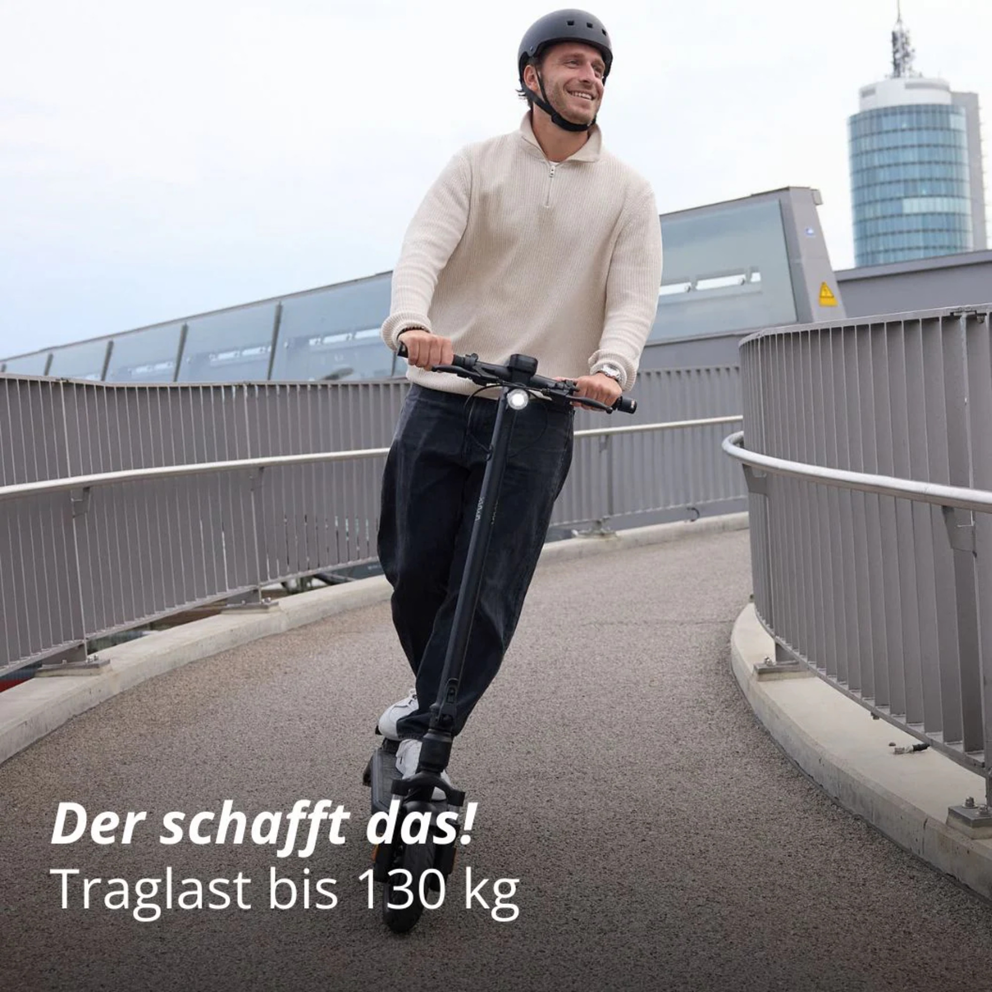 Ein Mann mit Helm fährt mit einem E-Scooter auf einer Brücke. Text: Der schafft das! Traglast bis 130 kg.