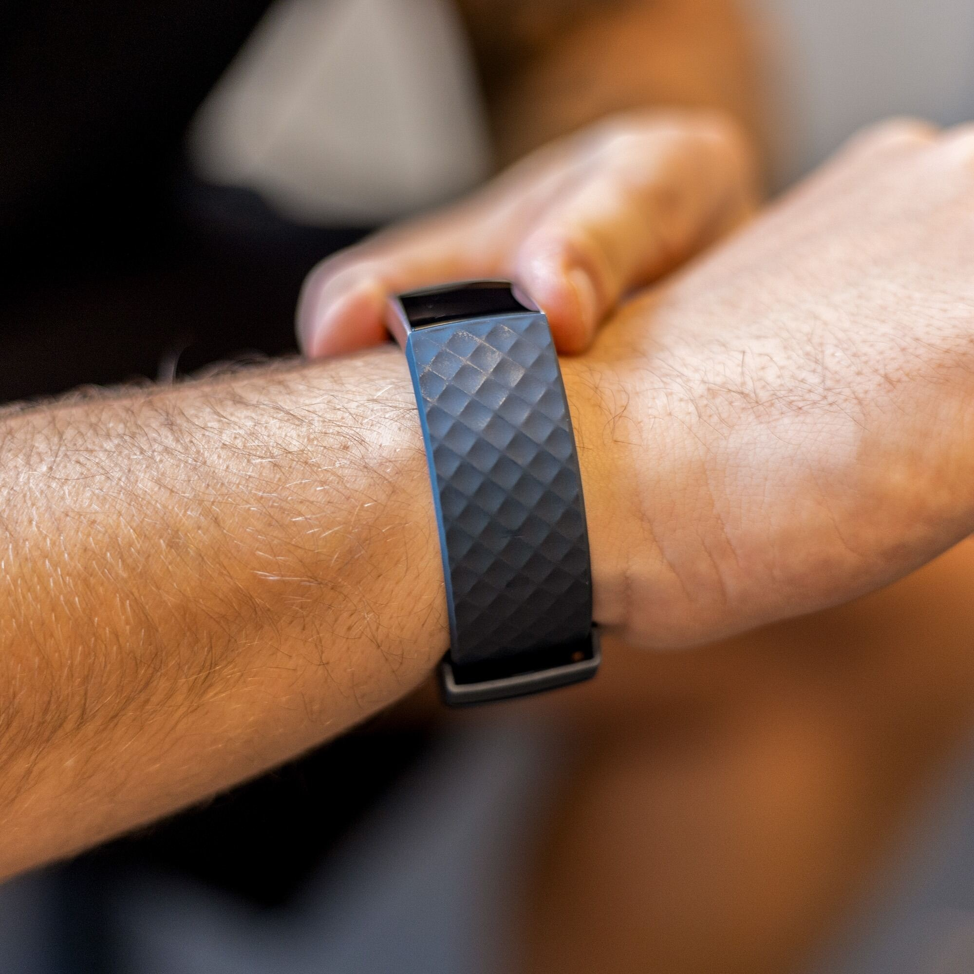 Ein Handgelenk mit einem schwarzen Smartband. Das Band hat ein Diamantmuster. Die Person trägt ein schwarzes Hemd.